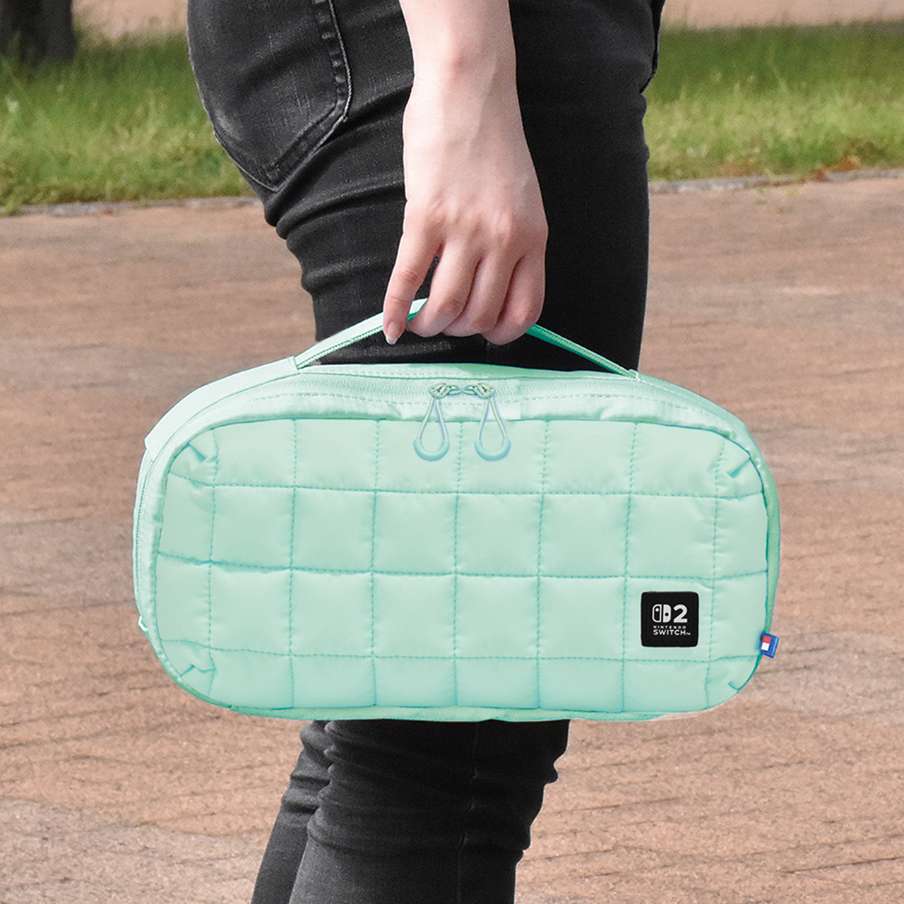 Une personne tient un étui matelassé vert menthe. L'étui a une poignée et des fermetures éclair. Un pantalon noir est visible, ainsi qu'un trottoir et de l'herbe.