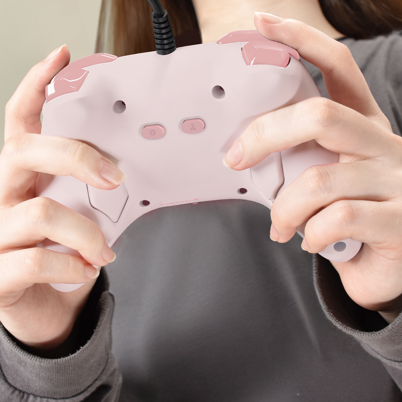 Une personne tient une manette Nintendo Switch rose et blanche. La manette est connectée par un câble noir. La personne porte un haut gris.