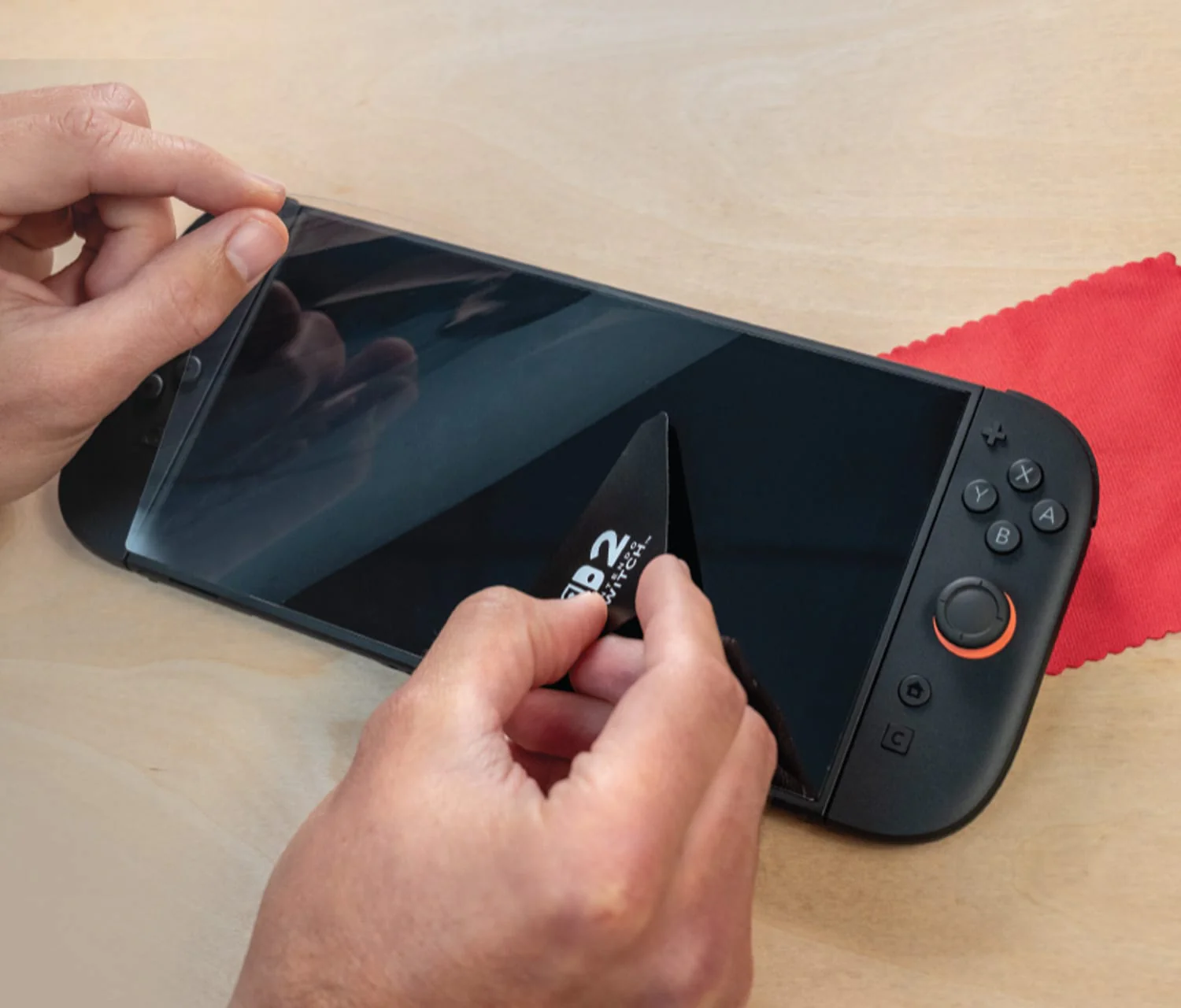Mains appliquant un protecteur d'écran sur une Nintendo Switch noire. Un chiffon rouge en arrière-plan. L'appareil est sur une surface en bois clair.