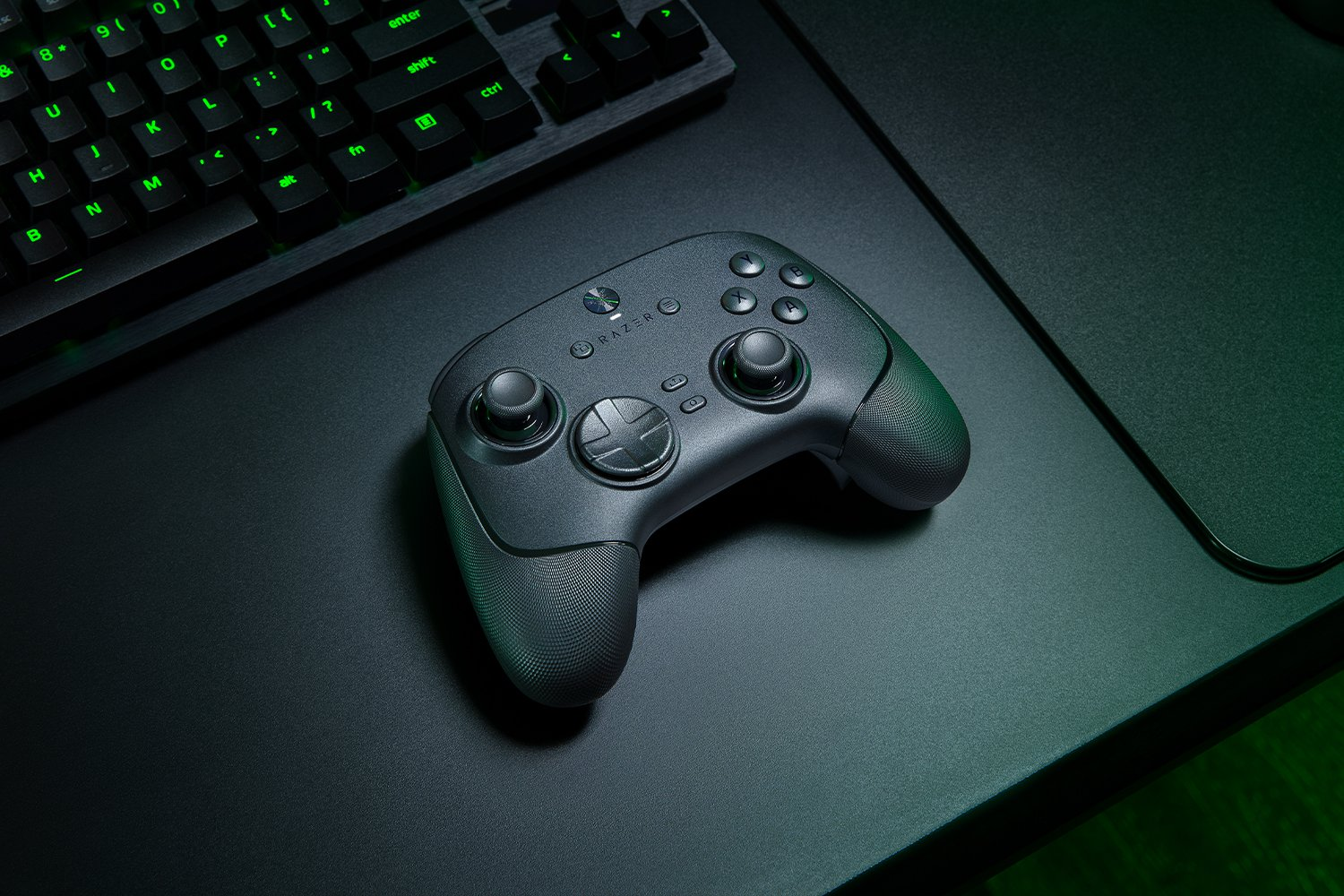 Ein schwarzes Razer-Gamepad auf einem schwarzen Schreibtisch neben einer Tastatur. Grüne Lichter sind auf der Tastatur sichtbar.