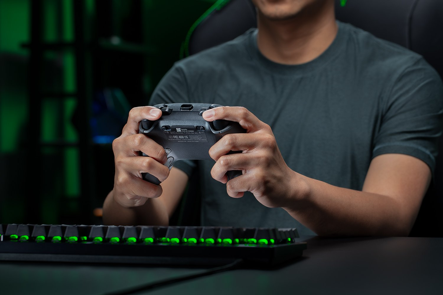 Eine Person hält einen grauen Gamecontroller. Im Vordergrund befindet sich eine Tastatur mit grün leuchtenden Tasten. Der Hintergrund ist dunkel und grün.