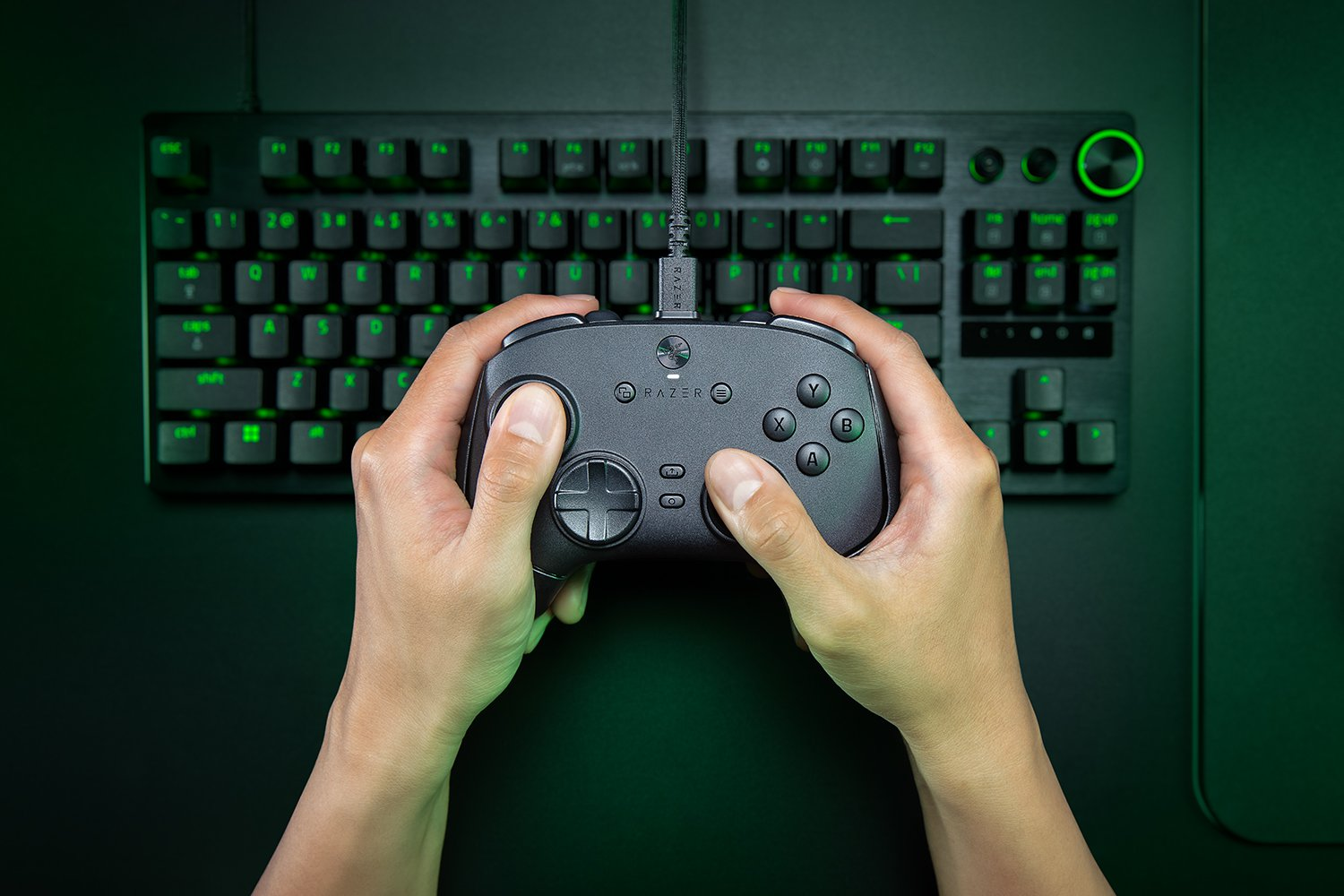 Mani che tengono un controller di gioco Razer, con una tastiera sullo sfondo. La tastiera e il controller hanno luci verdi.