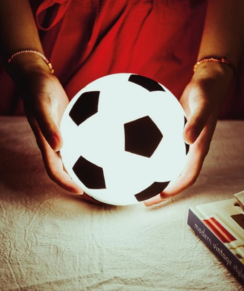 Dłonie trzymają świecącą piłkę nożną z czarnymi pięciokątami. Czerwona koszula w tle. Książka leży na teksturowanej powierzchni.