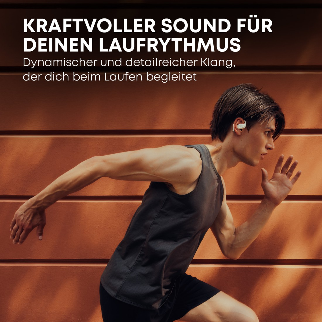 Ein Mann läuft mit Ohrhörern. Er trägt ein graues Tanktop und schwarze Shorts. Der Hintergrund ist eine braune Wand.