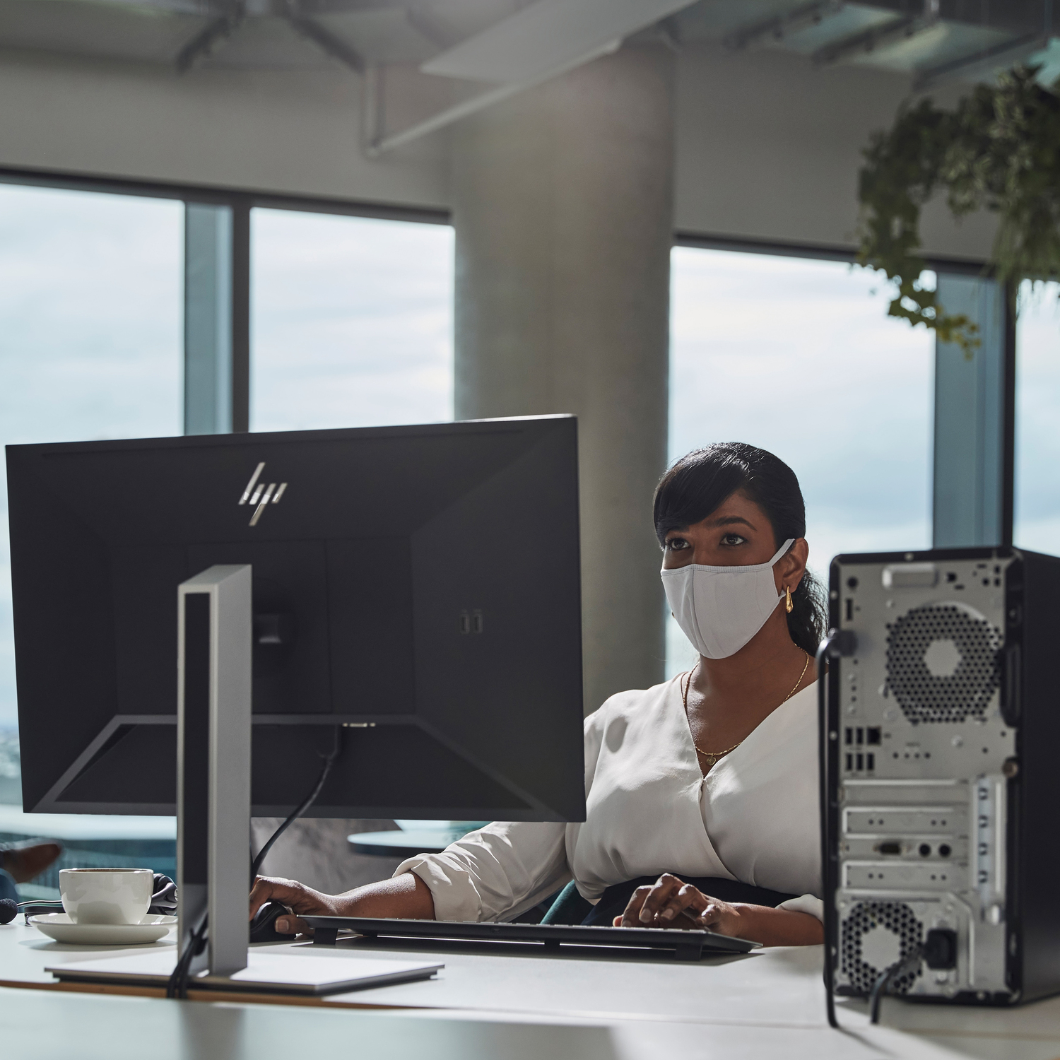 Una mujer con mascarilla trabaja en una computadora en una oficina. Se ven una taza de café, un monitor y una torre de computadora.