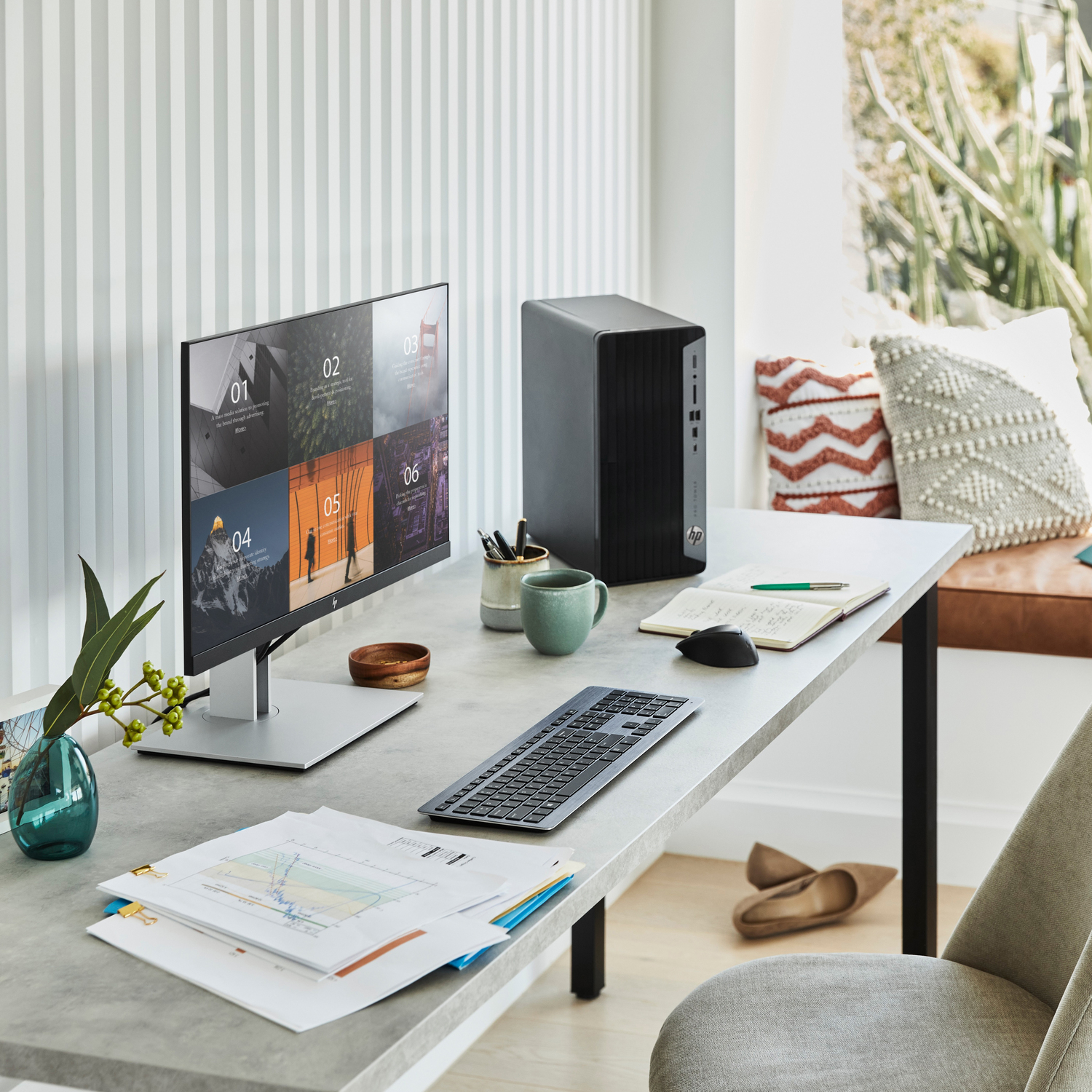 Un escritorio moderno. Un monitor de computadora muestra datos, con teclado y ratón. Hay una taza, un cuaderno y un jarrón con plantas.