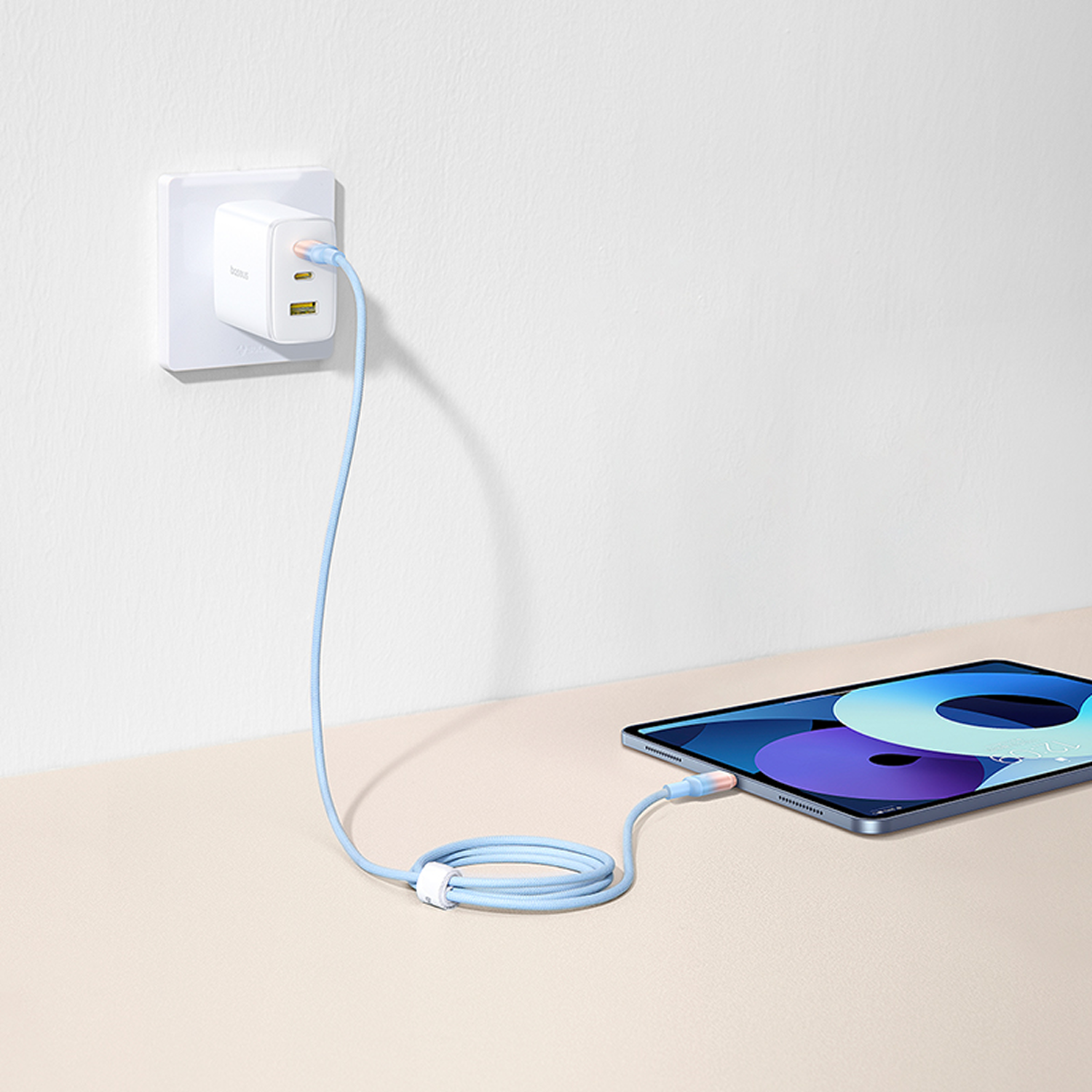 Un adaptador de corriente blanco enchufado a la pared. Un cable de carga azul conectado a una tableta sobre una superficie beige claro.