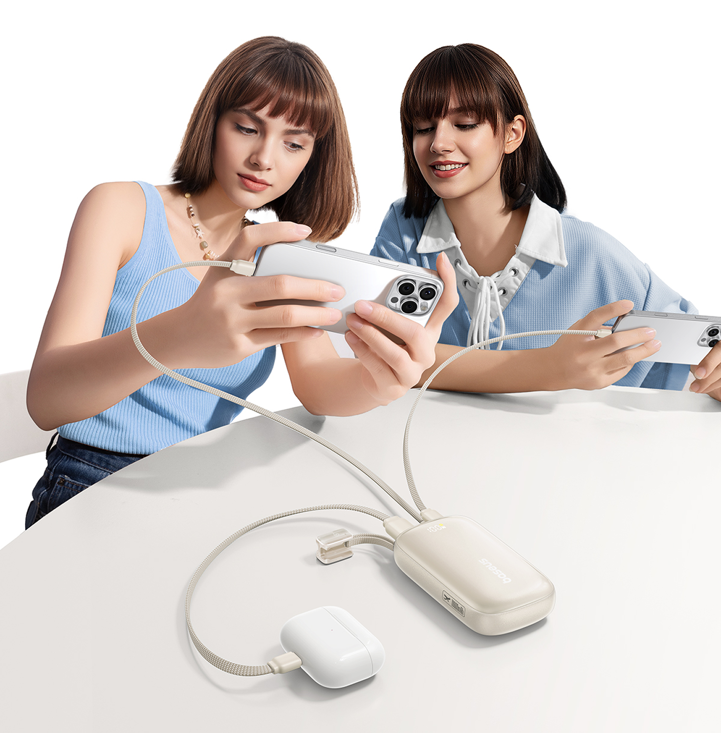 Dos mujeres sostienen teléfonos, conectados a un banco de energía y un estuche de auriculares. Están sentadas en una mesa blanca. La escena está bien iluminada.