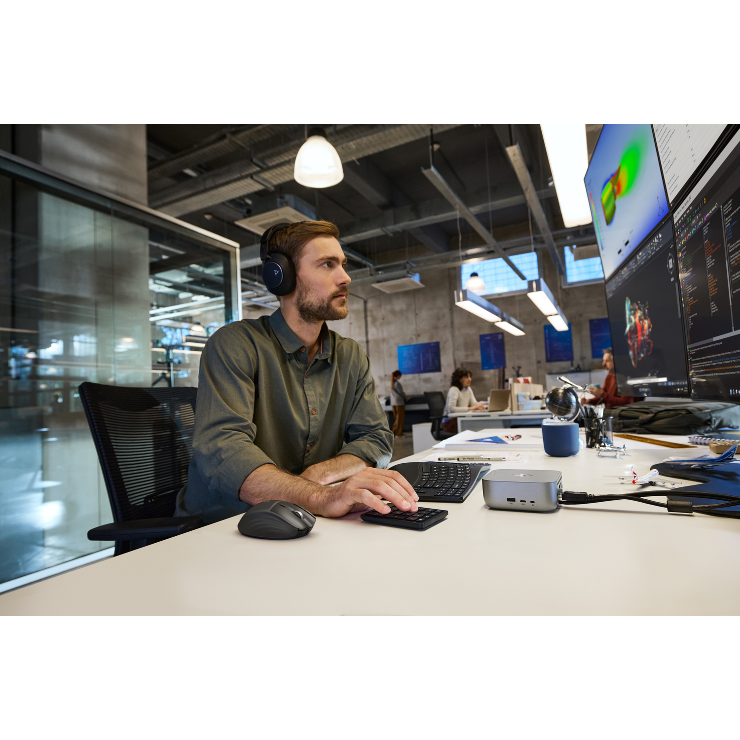 A man wearing headphones works at a desk with two monitors. He types on a keyboard and calculator. Other people are in the background.