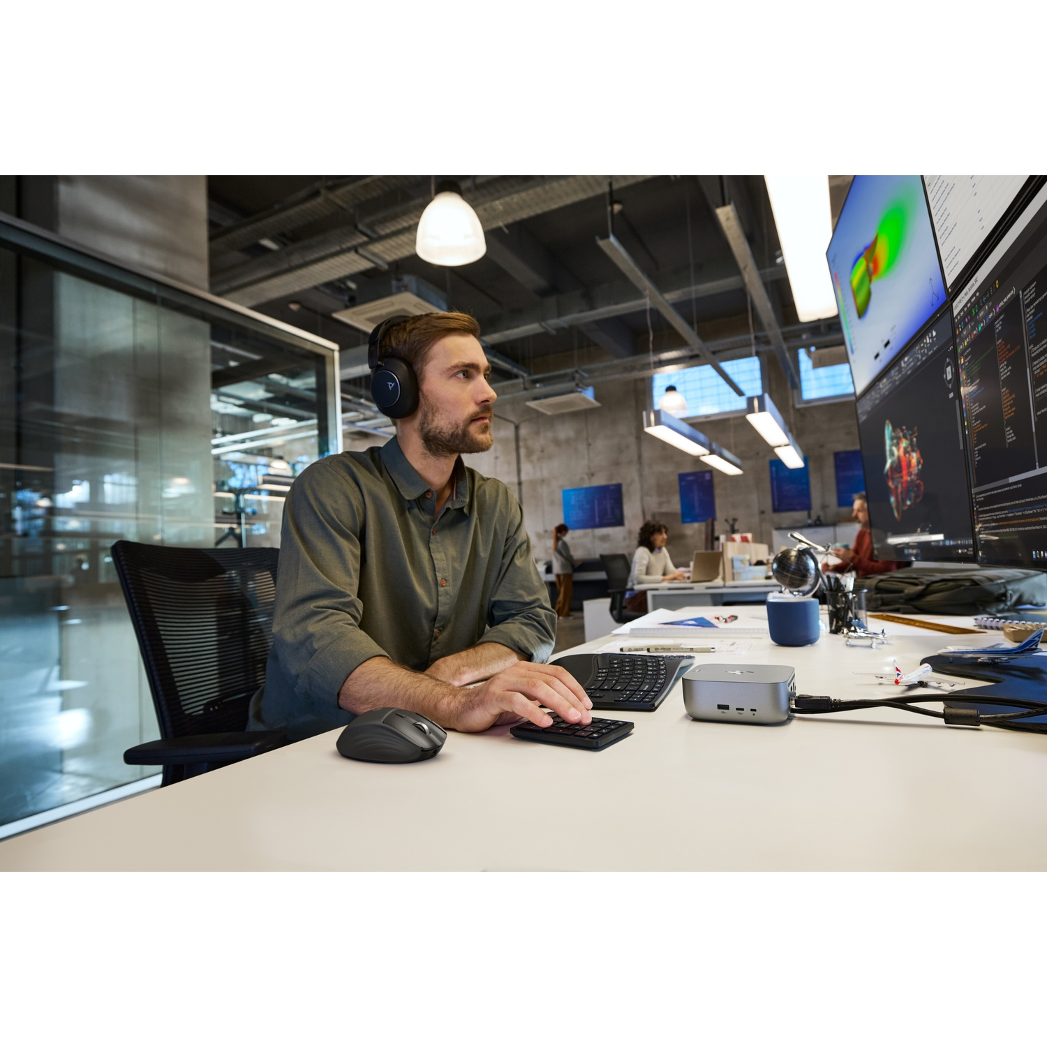 Un homme portant un casque travaille à un bureau avec deux écrans. Il utilise un clavier et une souris. D'autres personnes sont au fond.