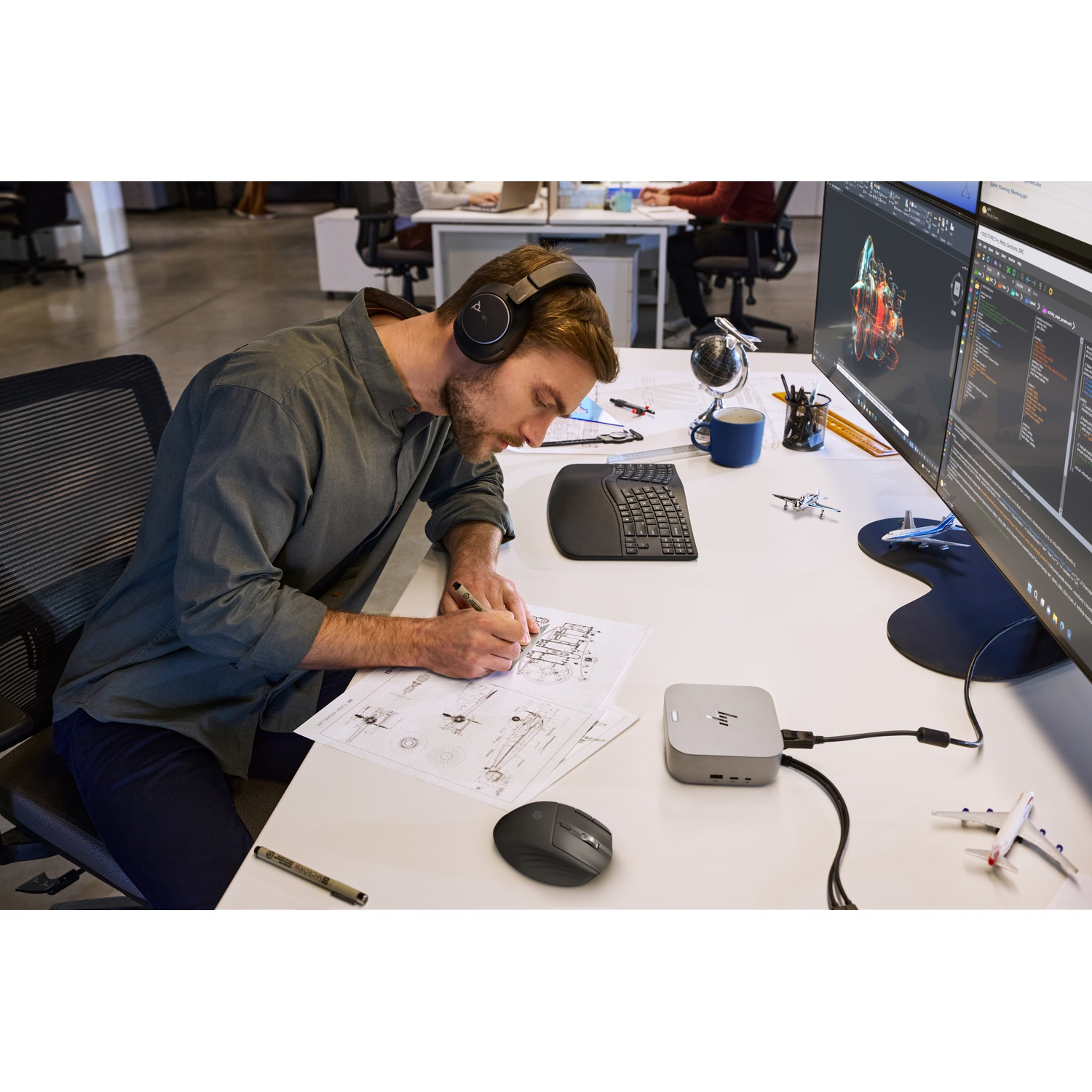 Un homme portant un casque dessine sur un plan de travail. Des écrans d'ordinateur et une souris sont visibles.