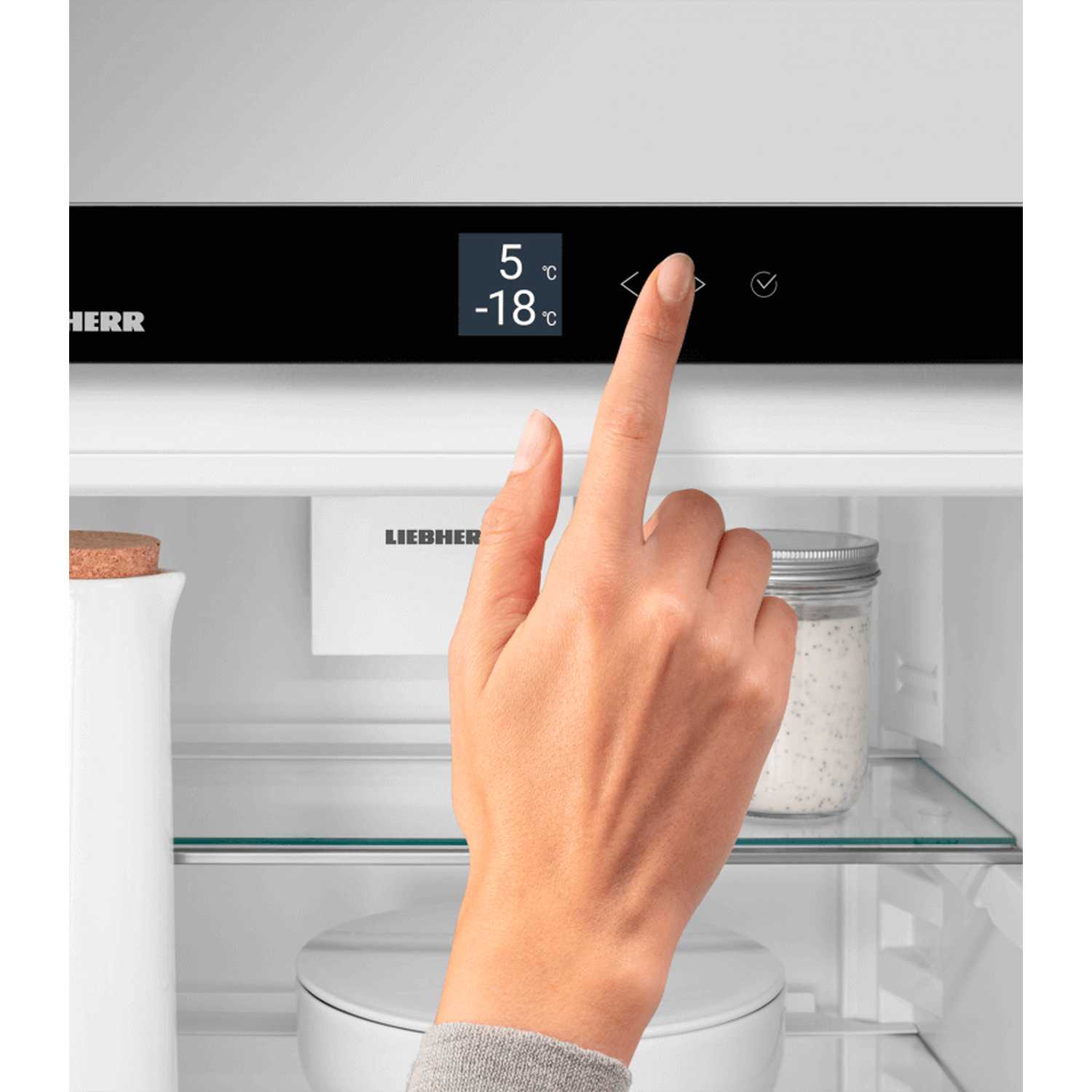 A hand touches a fridge control panel displaying temperatures. Inside, a jar and a white jug are visible.