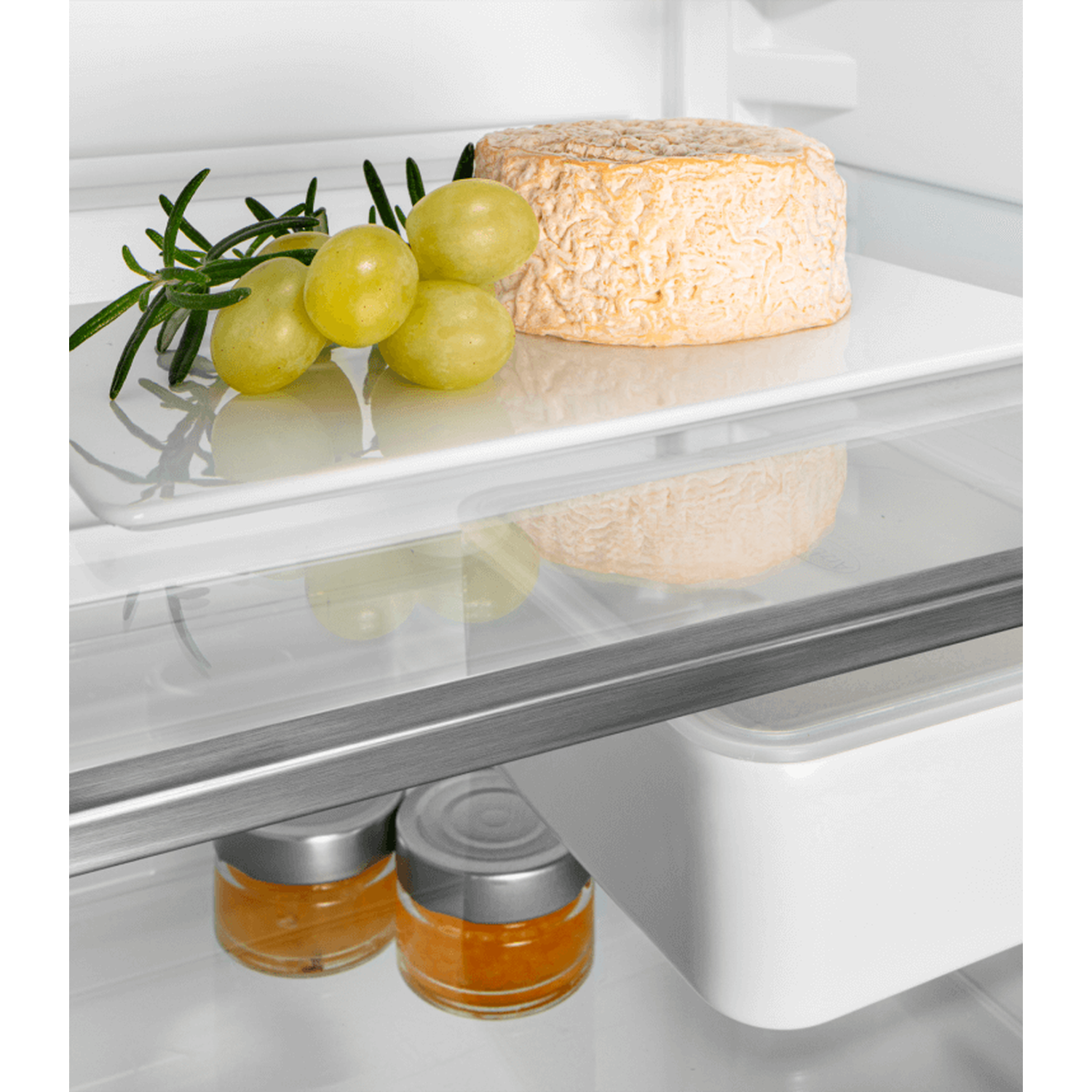 Inside a fridge: Cheese, green grapes, and rosemary on a shelf. Below, jars of orange jam are visible.