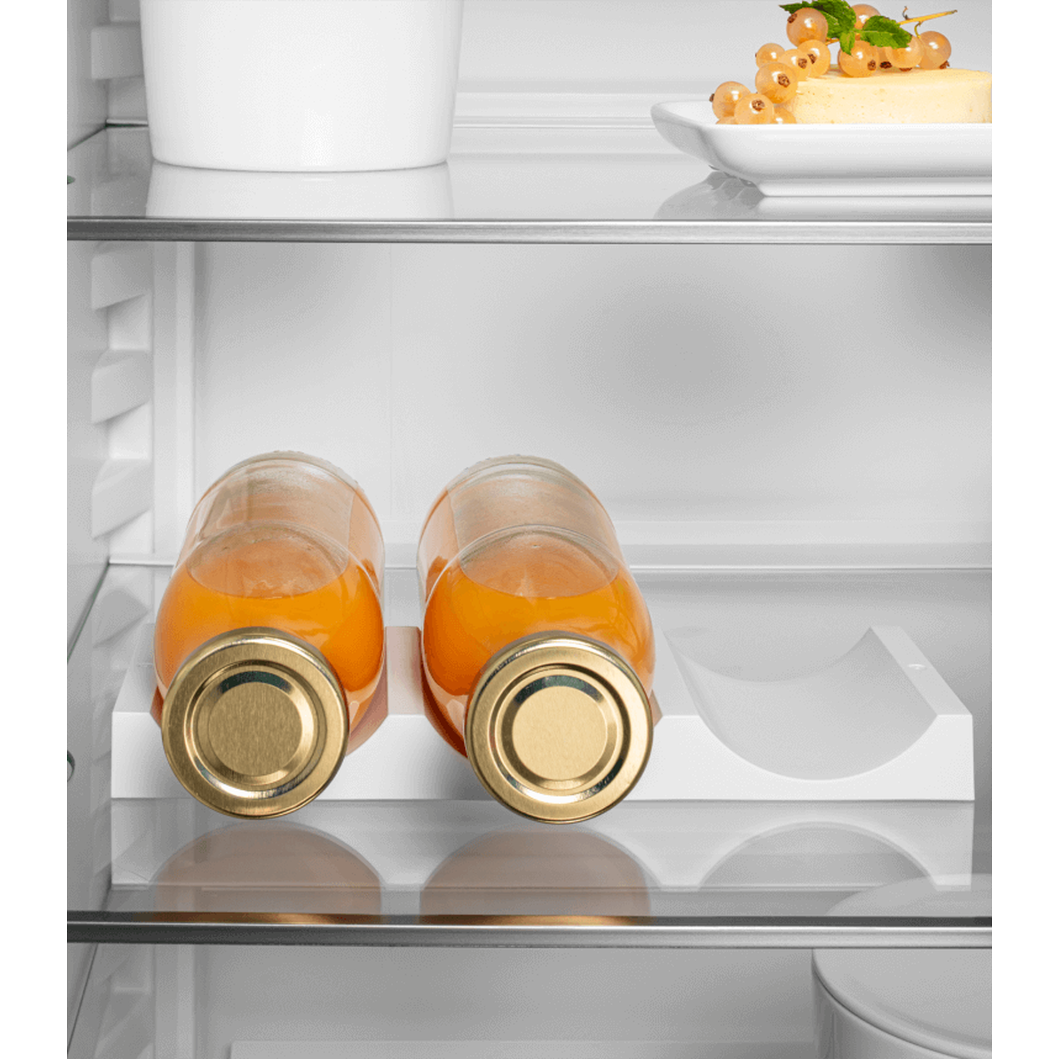 Inside a refrigerator, two bottles of orange juice sit on a white shelf. Other shelves hold food and a container.