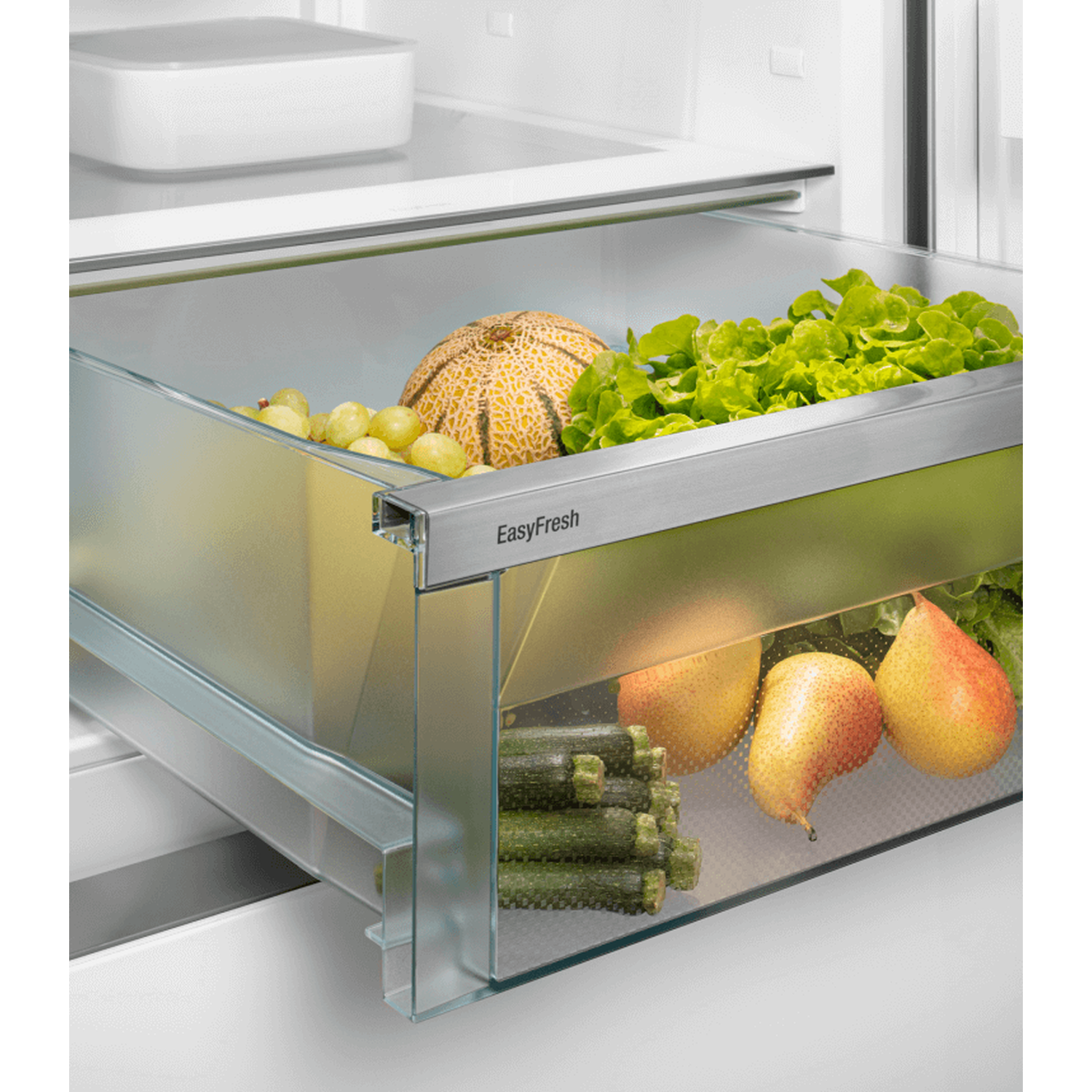 Inside a refrigerator drawer, there is a melon, grapes, lettuce, pears and zucchini. The drawer is made of glass.