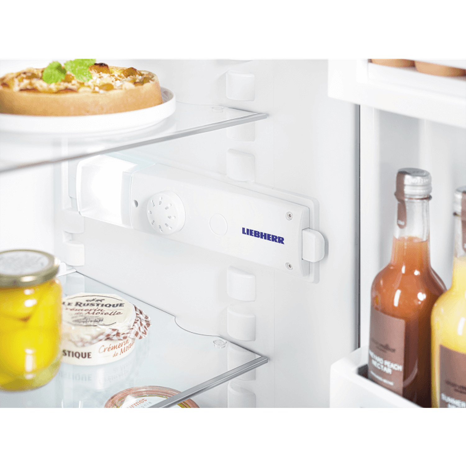 Inside a refrigerator: pizza, cheese, jar, bottles, and a control panel with the Liebherr logo. White interior.