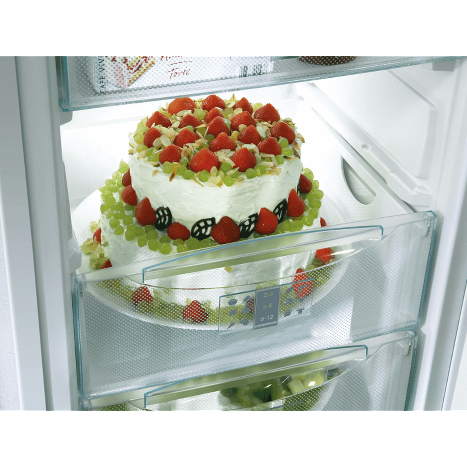 A cake with strawberries, grapes and almonds in a fridge. The cake is on a white plate. Inside the fridge are shelves.