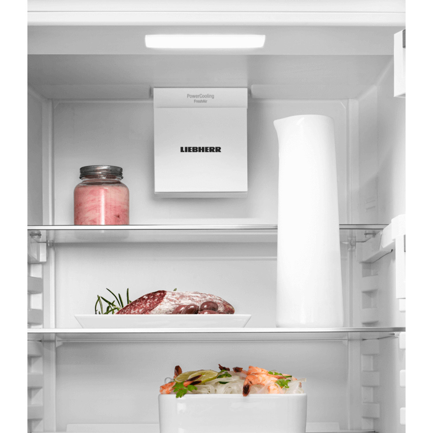 Inside a fridge: jar of pink food, white pitcher, plate with meat and herbs, and bowl of shrimp noodles.