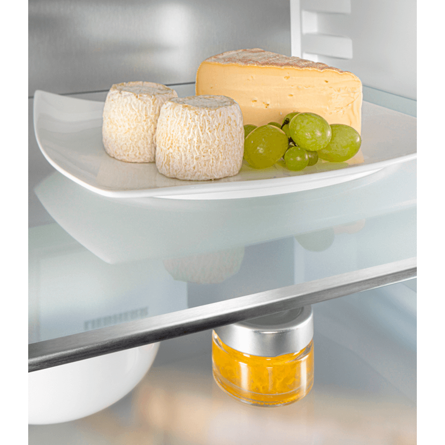 Inside a fridge: white plate with cheese and grapes. Below, a jar of yellow substance. Background is the fridge interior.
