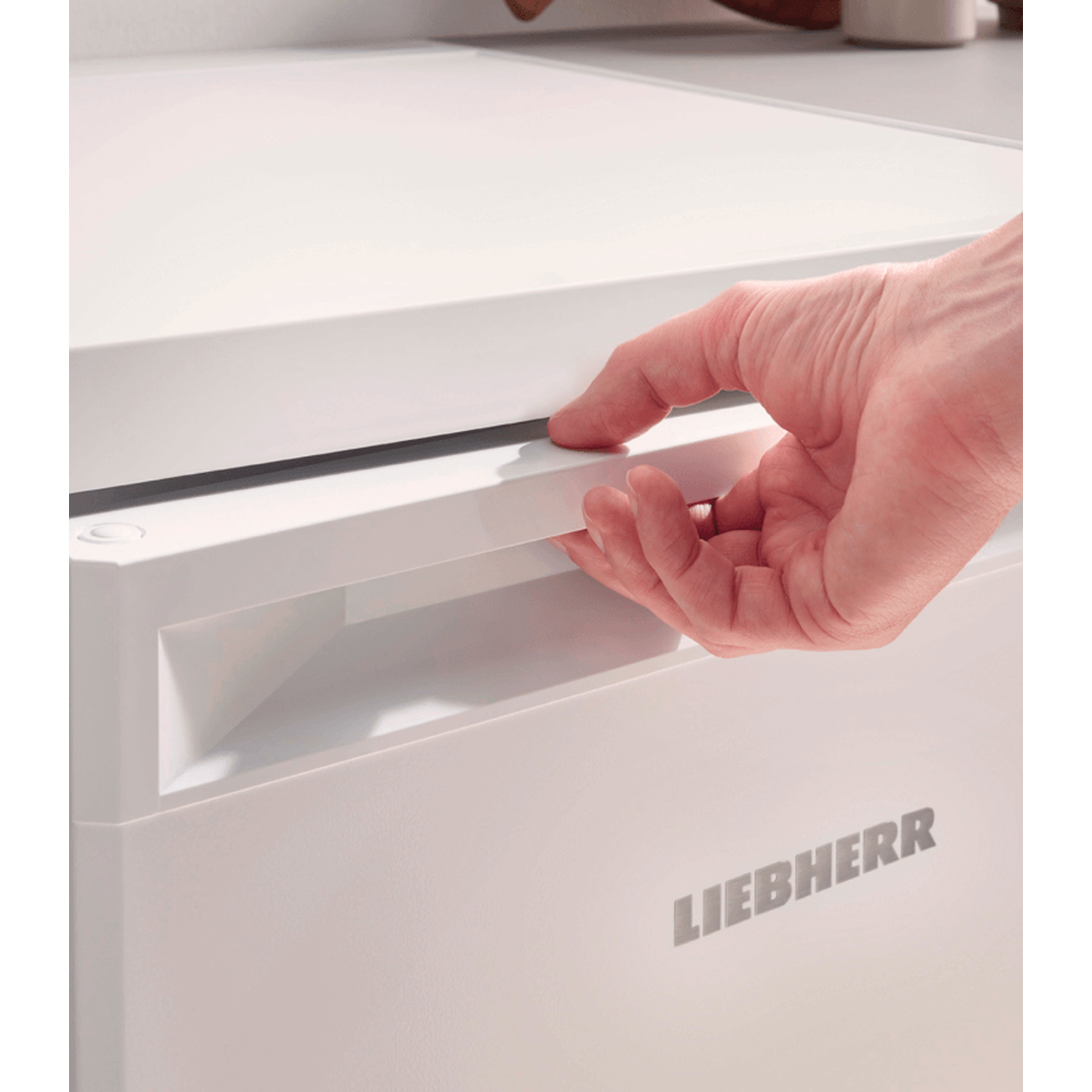 A hand opens a white refrigerator drawer. The brand name LIEBHERR is visible. The scene is indoors with bright lighting.