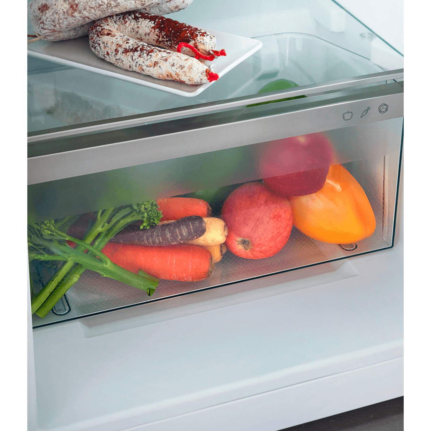 Inside a refrigerator: sausages on top shelf, below apples, carrots, broccoli, and a yellow pepper.