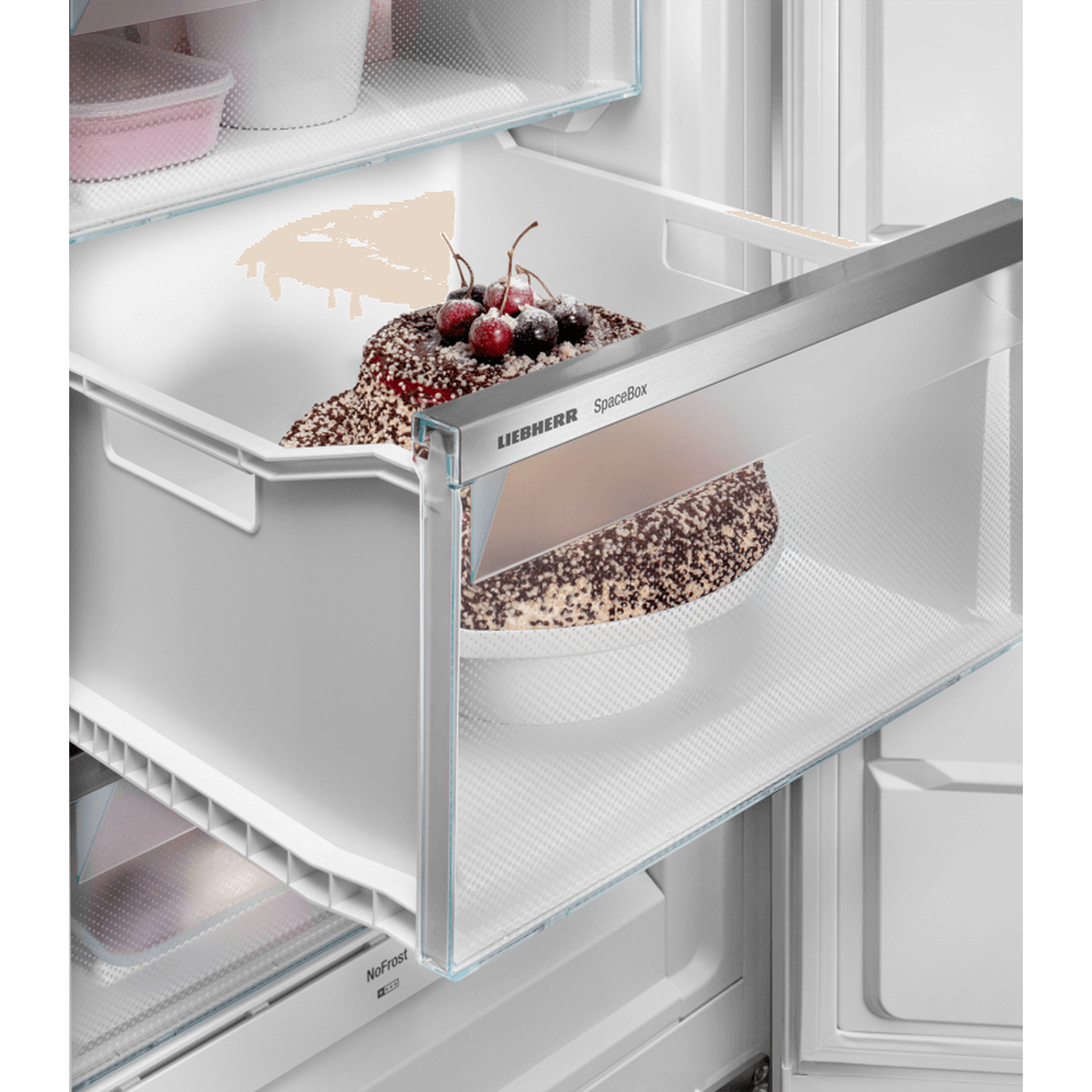 Inside a fridge drawer, a chocolate cake with cherries sits. The drawer is open, revealing a white interior. Other items are visible.