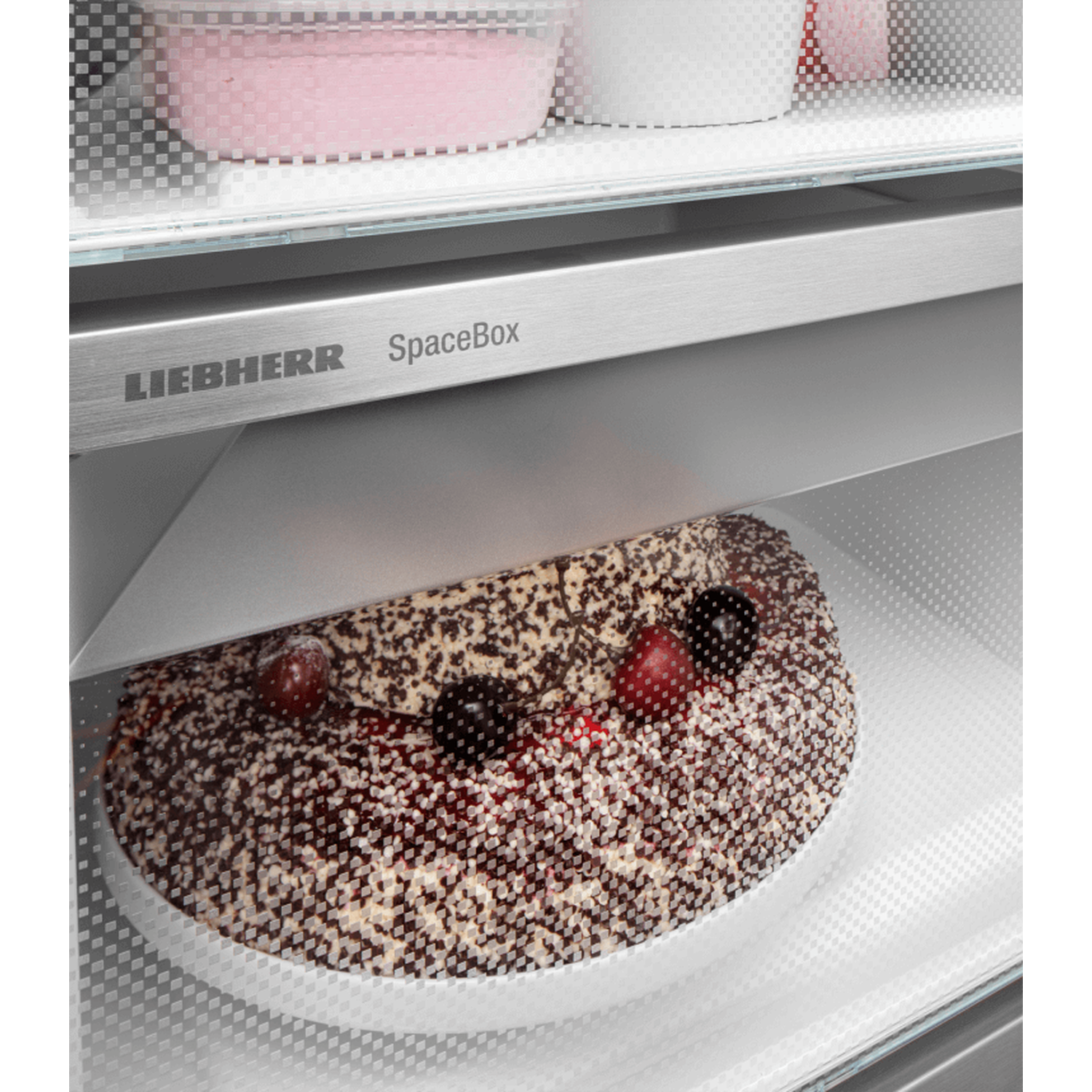 Inside a fridge: a cake with cherries, in front of the Liebherr SpaceBox sign. Pink and white containers are on the shelf above.