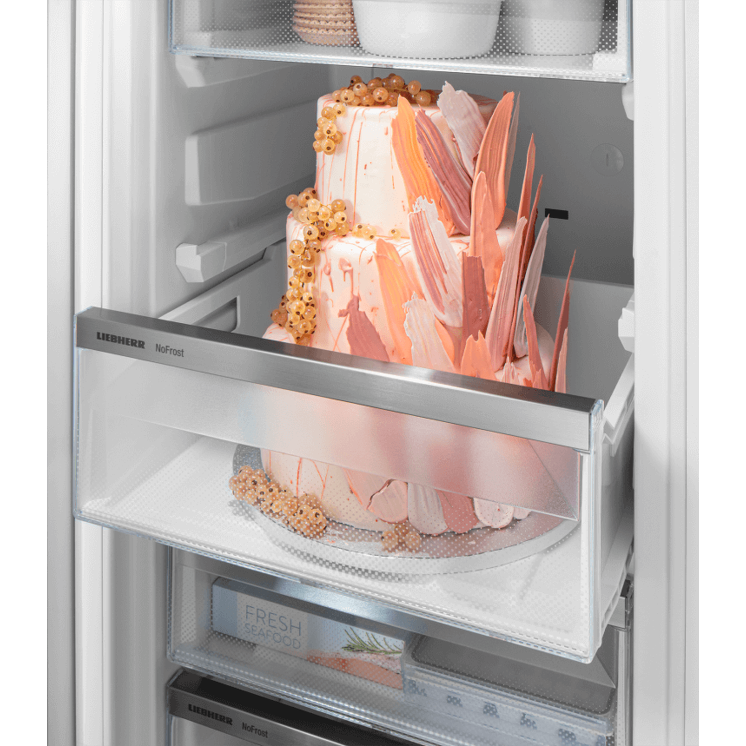 A two-tiered cake with pink decorations and berries is in a refrigerator. Below, there's another cake. Fresh seafood box visible.