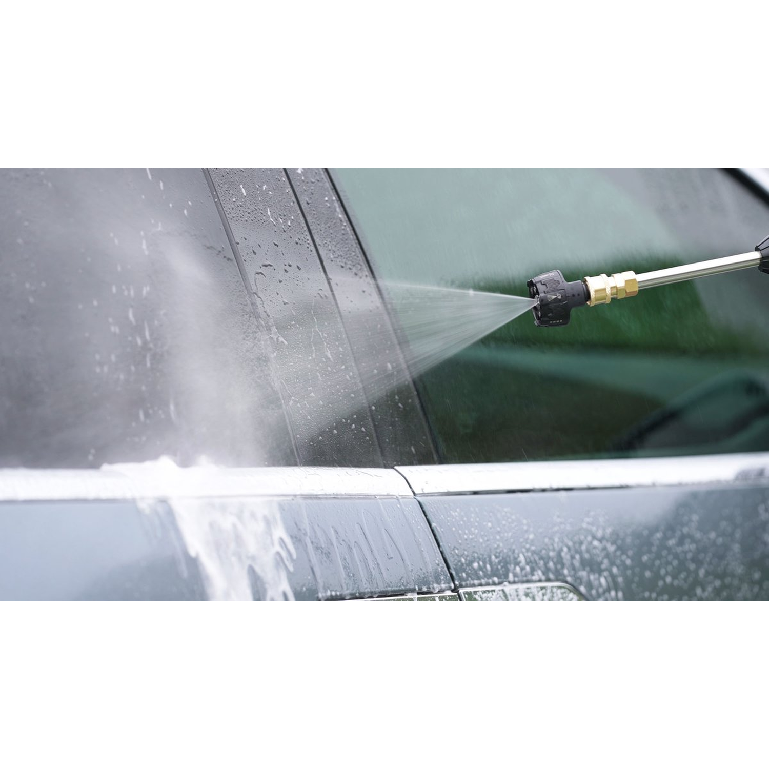 A car is being washed with a pressure washer. Water sprays onto the car's window and body, creating foam and water droplets.