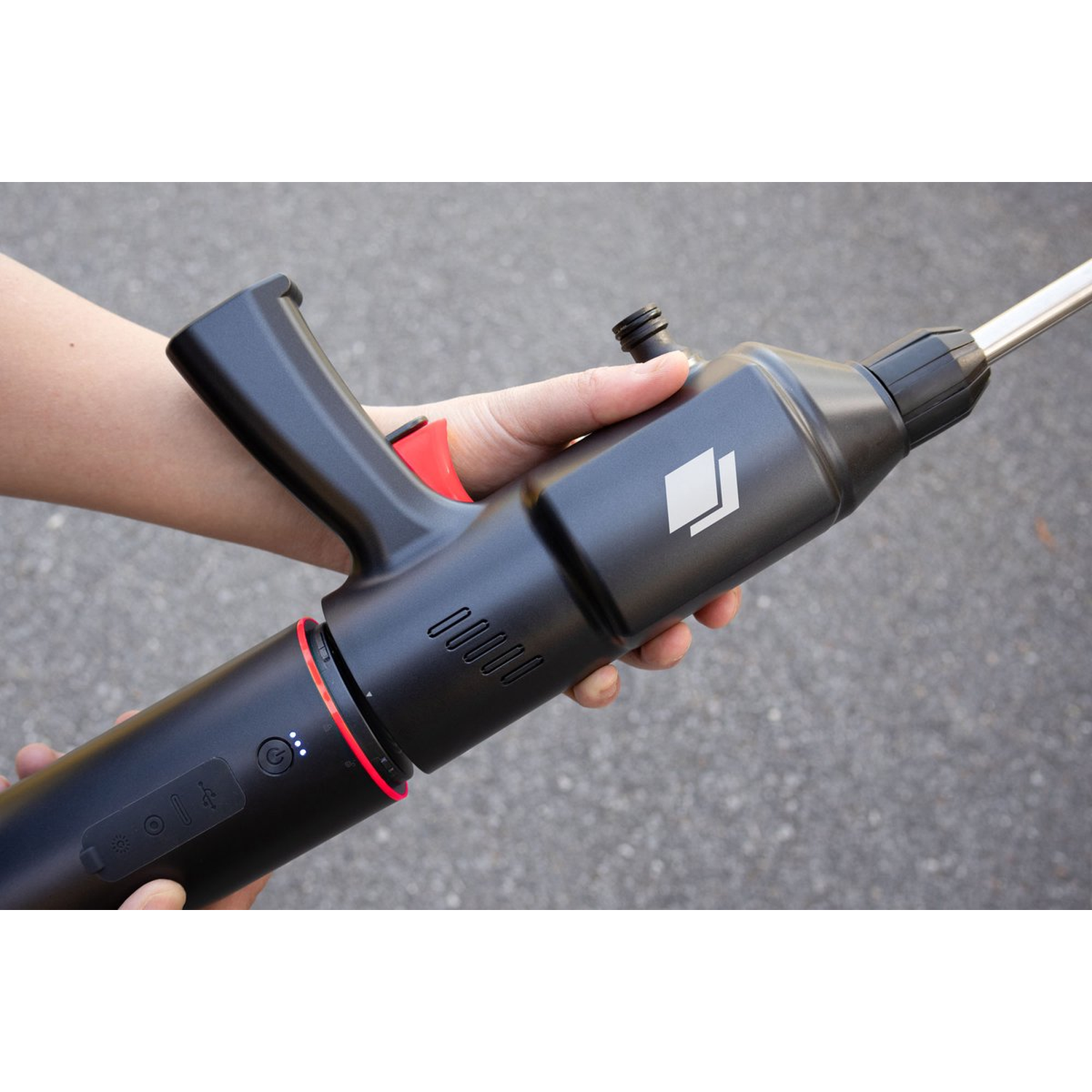 A person holds a black and red device. The device has a handle, a red button, and a silver nozzle. The background is gray asphalt.