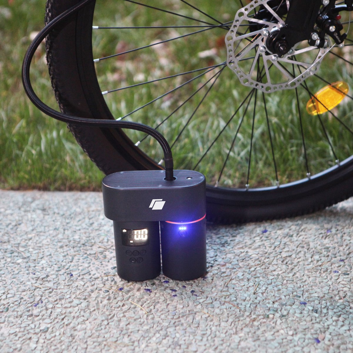A black bicycle tire with a disc brake. A black air pump with a display is connected to the tire with a hose. The pump is on a stone surface.