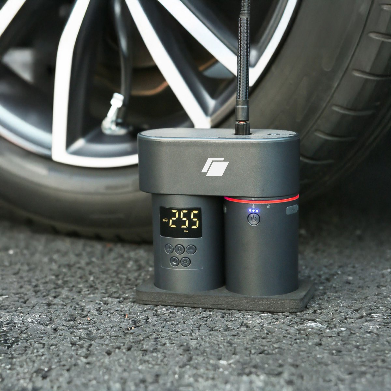 A black and gray tire inflator is on asphalt, next to a car tire. The inflator has a digital display and buttons.