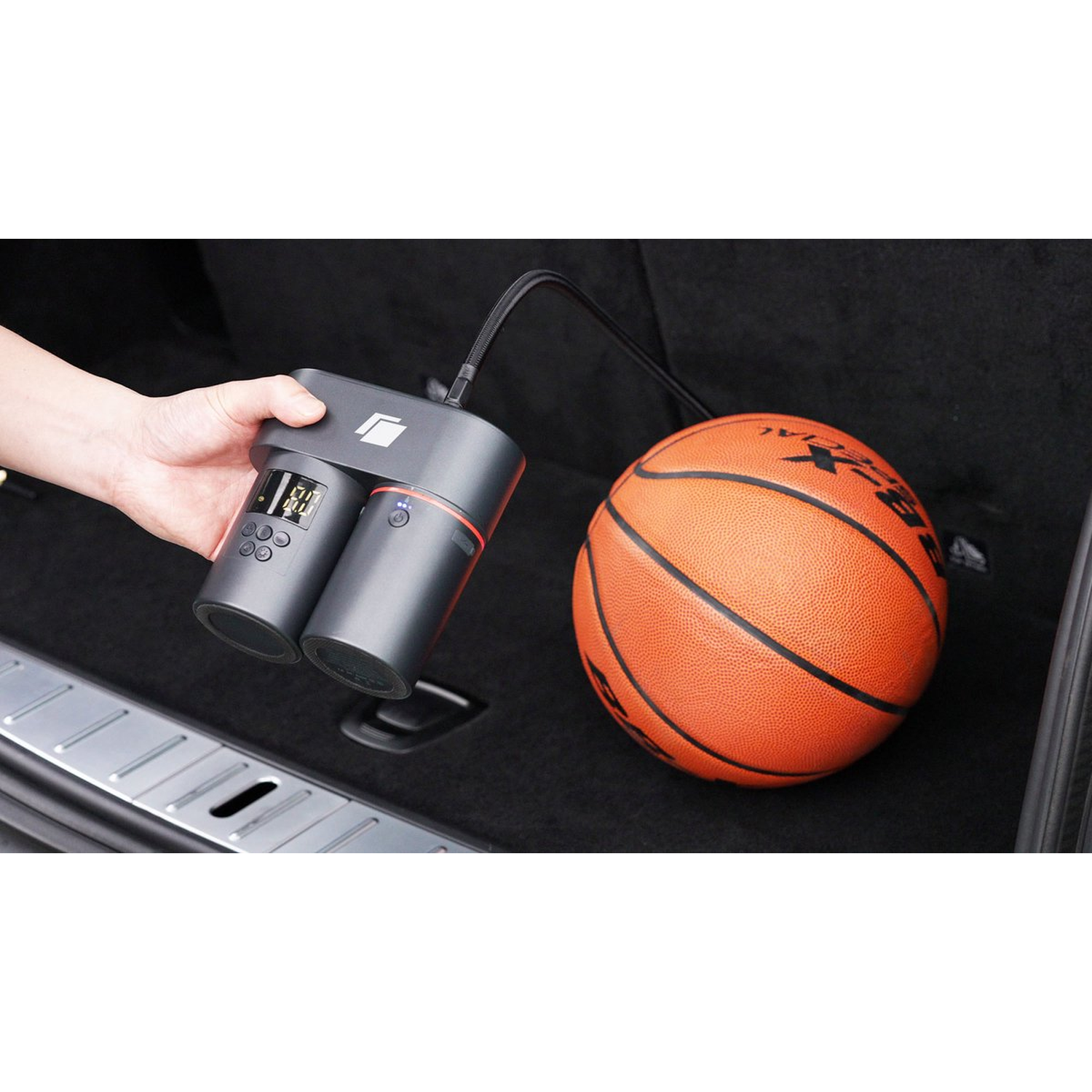 A person holding an air compressor connected to a basketball in a car trunk. The compressor is black and red. The ball is orange.