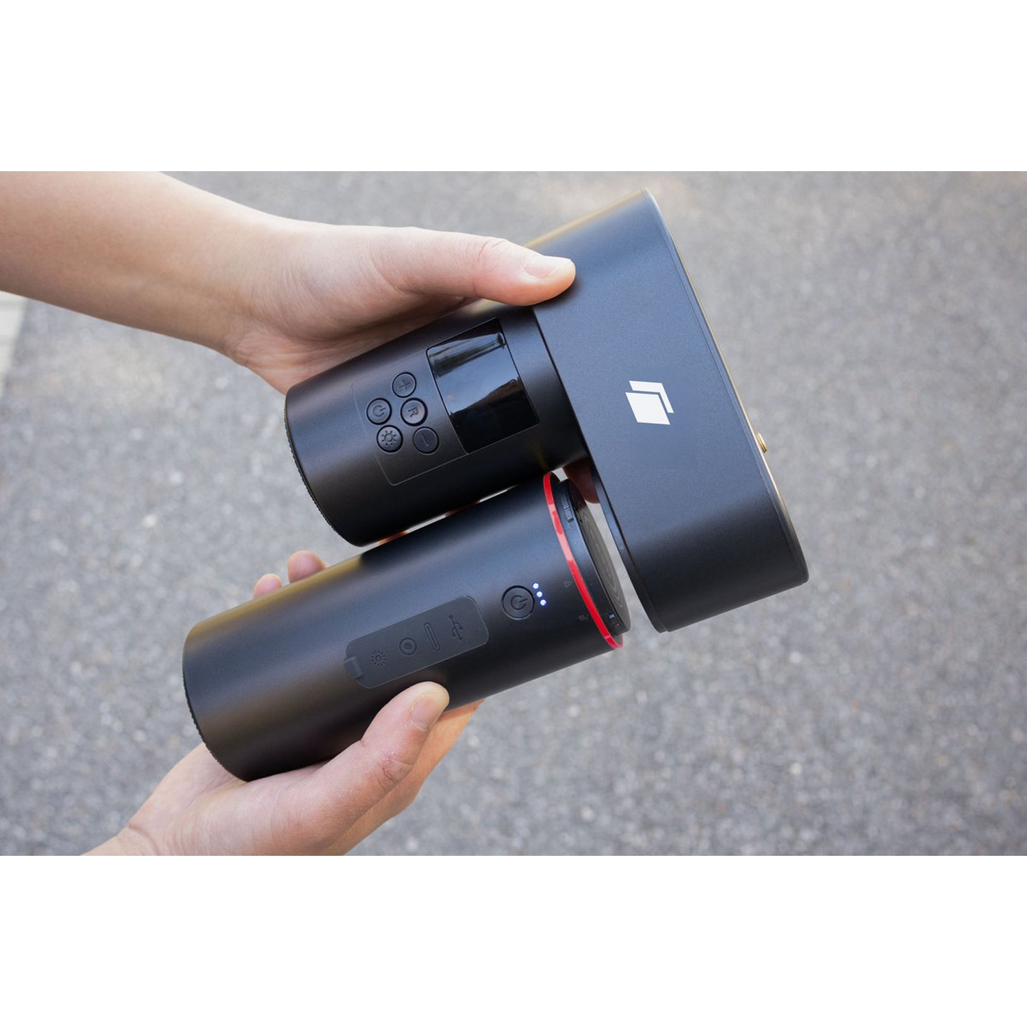 Hands holding a black device with buttons and a screen. The device is cylindrical with a red ring.