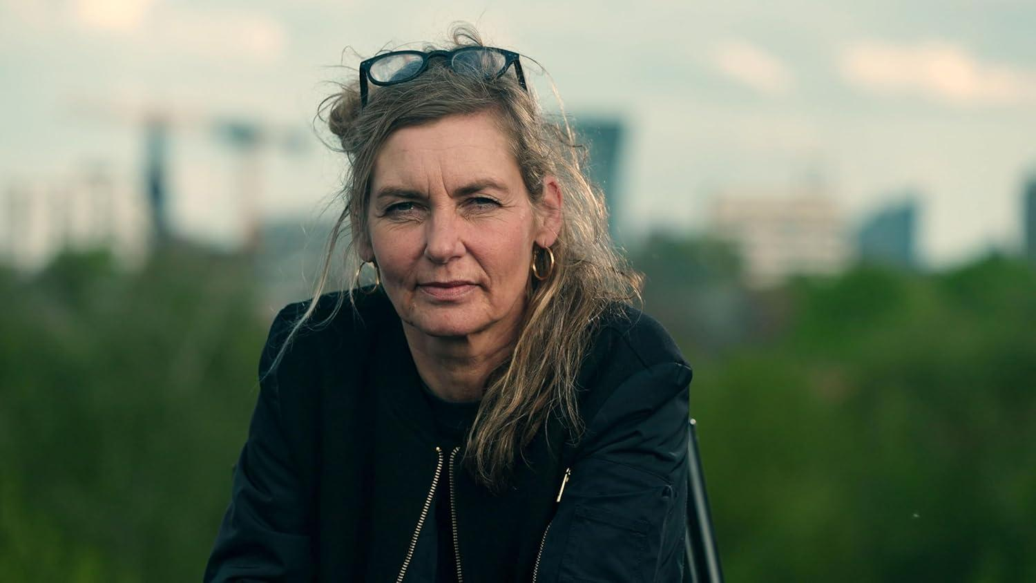Reife Frau mit Brille auf dem Kopf, blickt in die Kamera. Sie trägt eine schwarze Jacke, goldene Ohrringe und graue Haare. Stadt im Hintergrund.