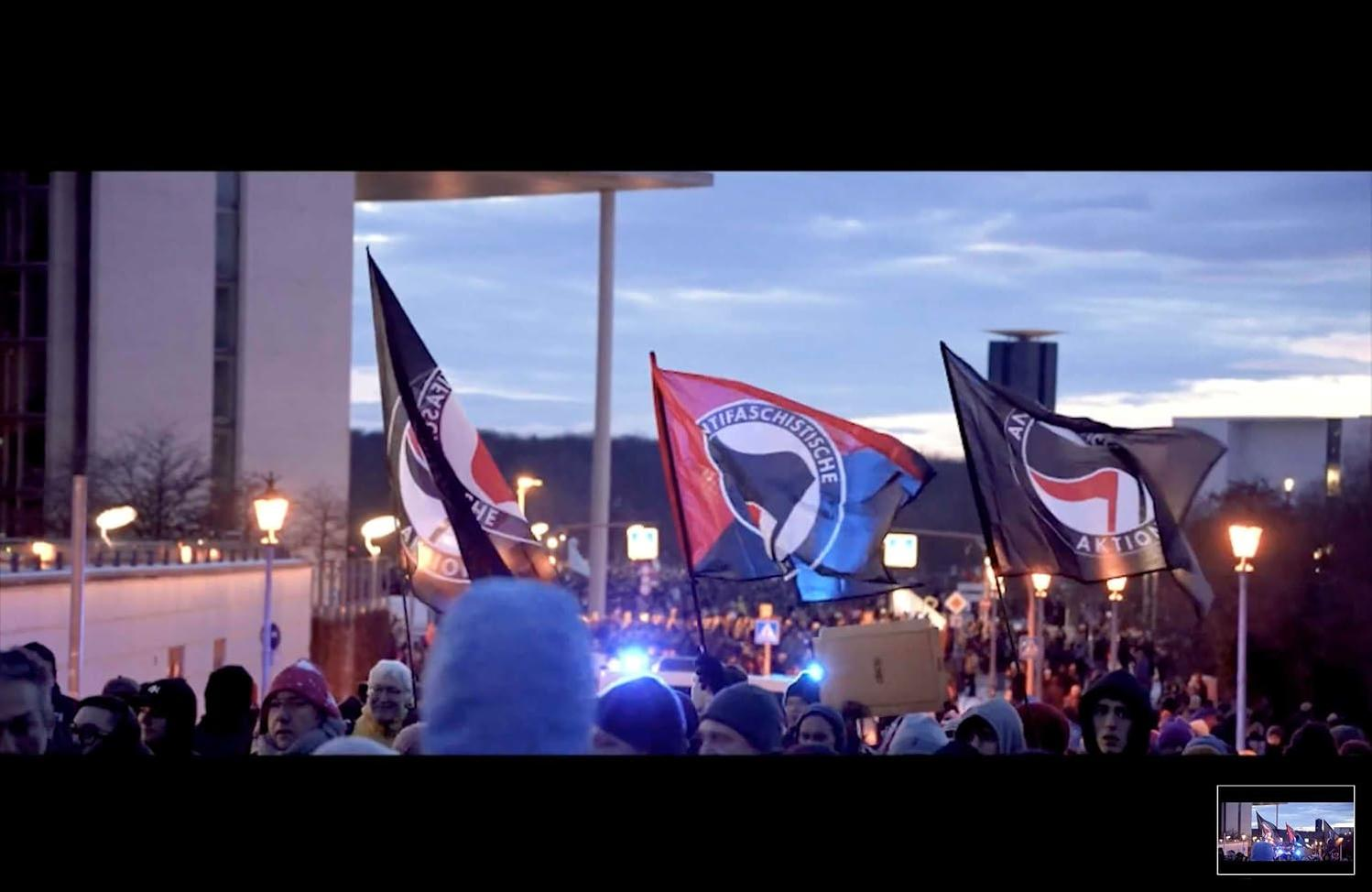 Demonstration mit Flaggen, darunter 'Antifaschistische Aktion'. Menschen im Vordergrund. Ein Gebäude ist im Hintergrund sichtbar.