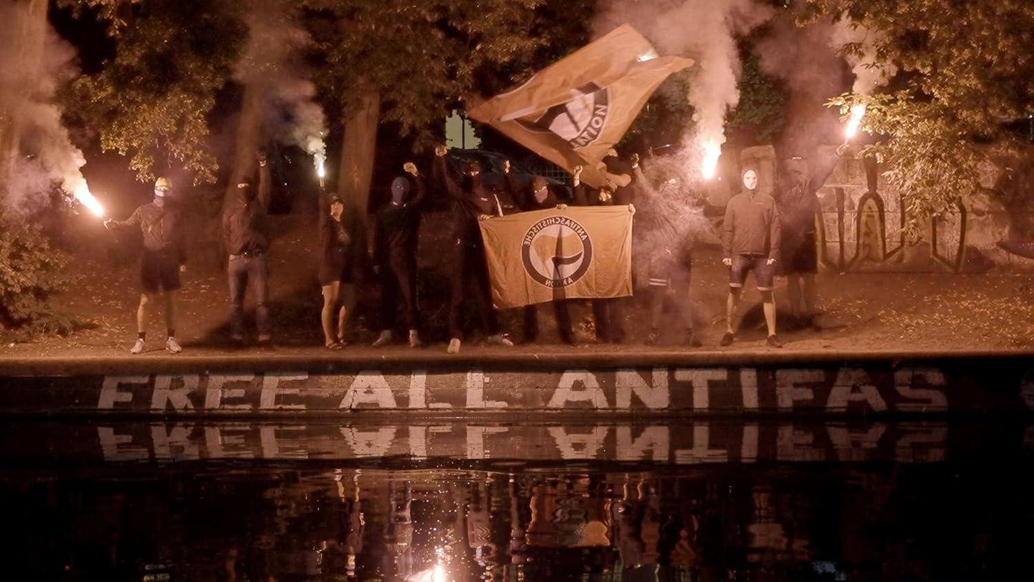 Gruppe von Personen mit Fahnen und Fackeln. Text 'FREE ALL ANTIFAS' ist sichtbar. Nachtszene mit Wasserspiegelung.