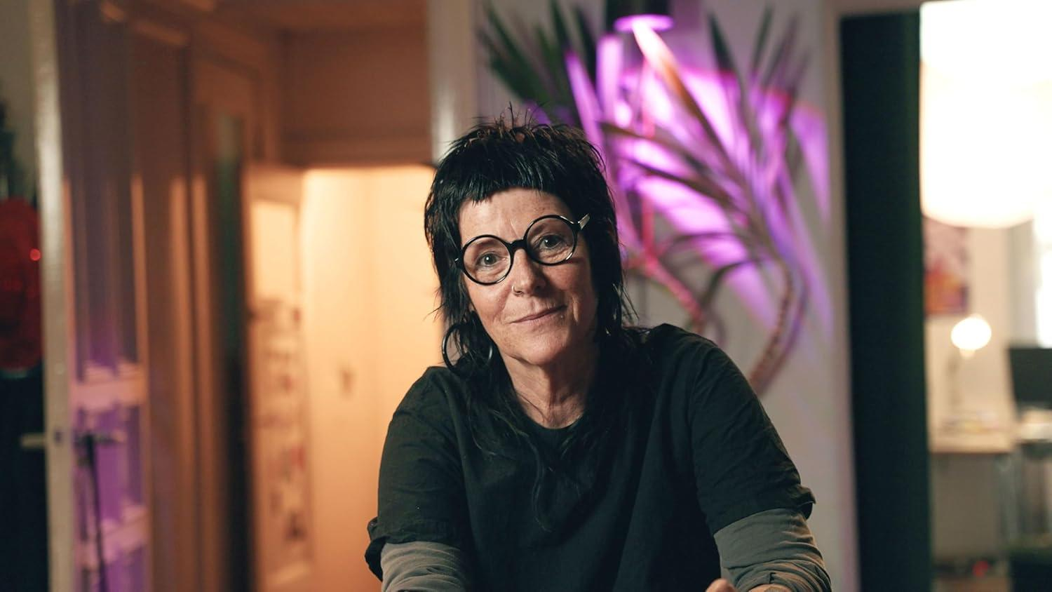 Eine Frau mit Brille und dunklem Haar lächelt. Sie trägt ein schwarzes Hemd in einem Raum mit einem Gemälde und Lichtern.
