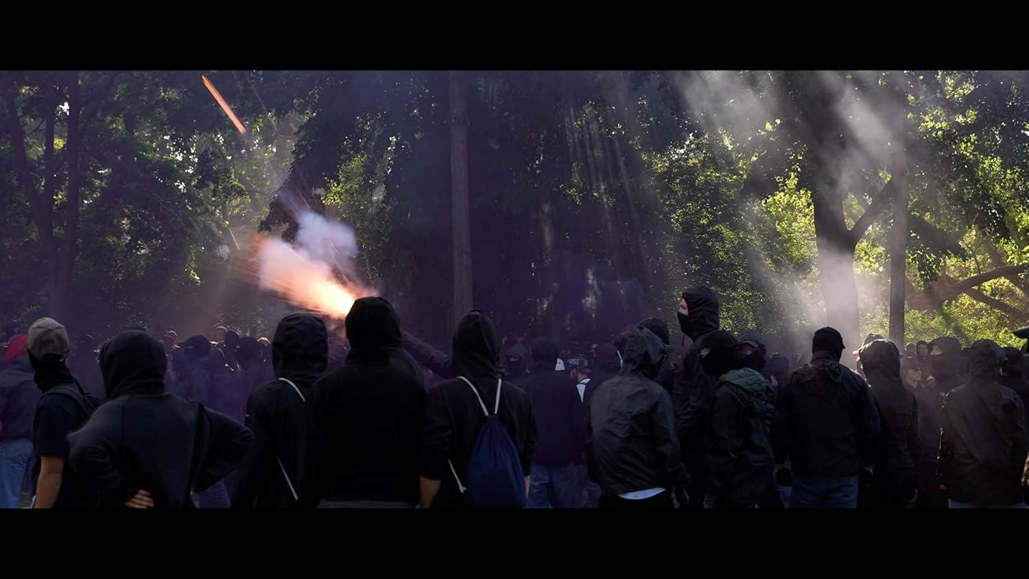 Eine Menge Menschen, viele in Schwarz, beobachten eine Explosion in einem Wald. Rauch und Lichtstrahlen sind sichtbar.