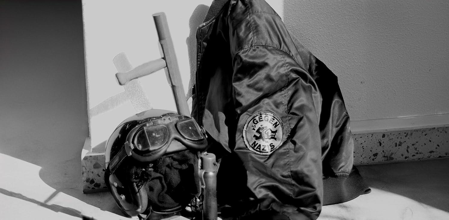 Schwarzer Helm mit Brille, ein Schlagstock und eine Jacke mit einem Aufnäher mit der Aufschrift "Gegen Nazis" lehnen an einer weißen Wand.