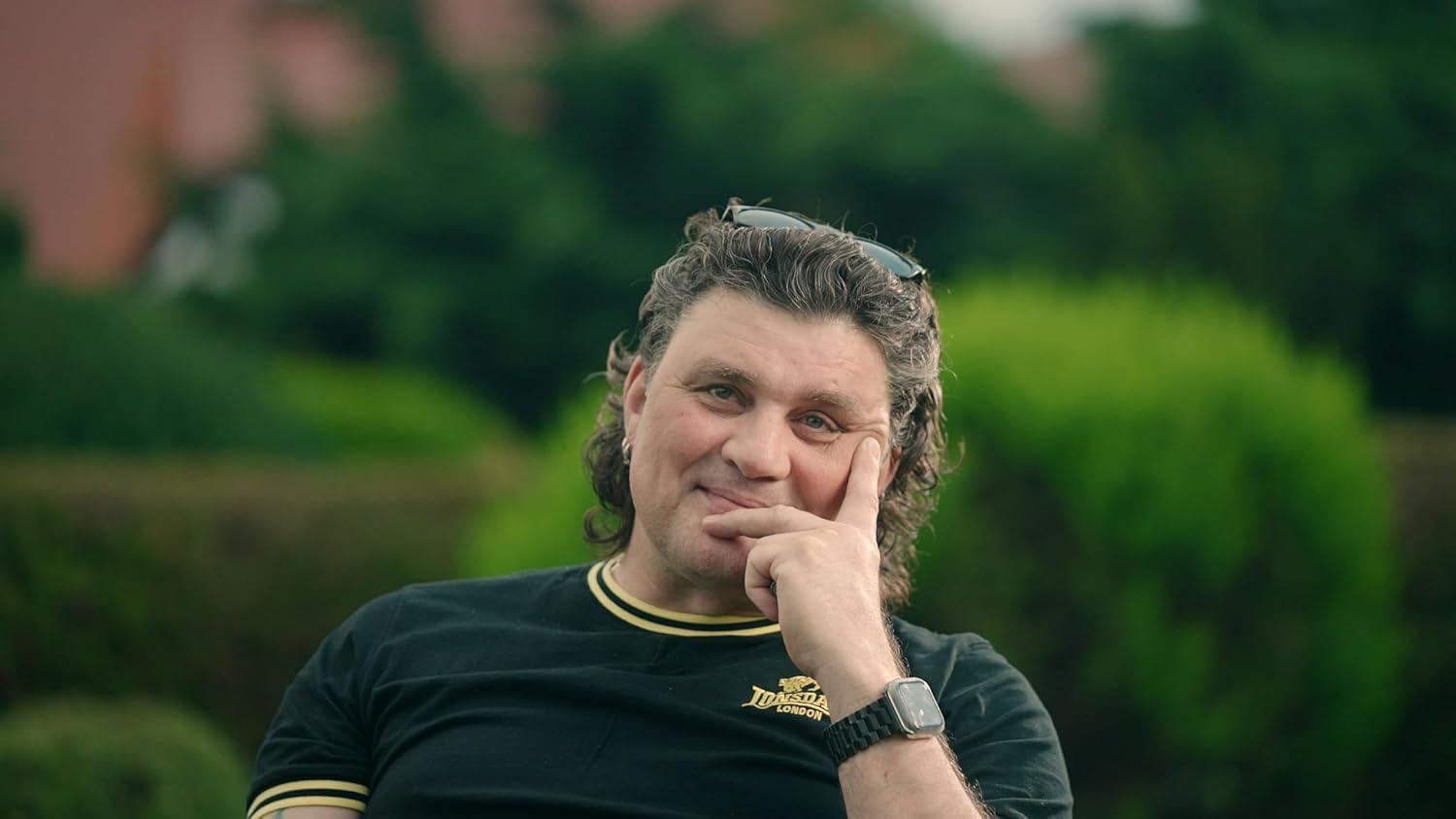 Ein Mann lächelt und stützt sein Kinn auf einen Finger. Er trägt ein schwarzes Hemd mit goldenen Rändern und eine Sonnenbrille auf dem Kopf, mit grünem Hintergrund.