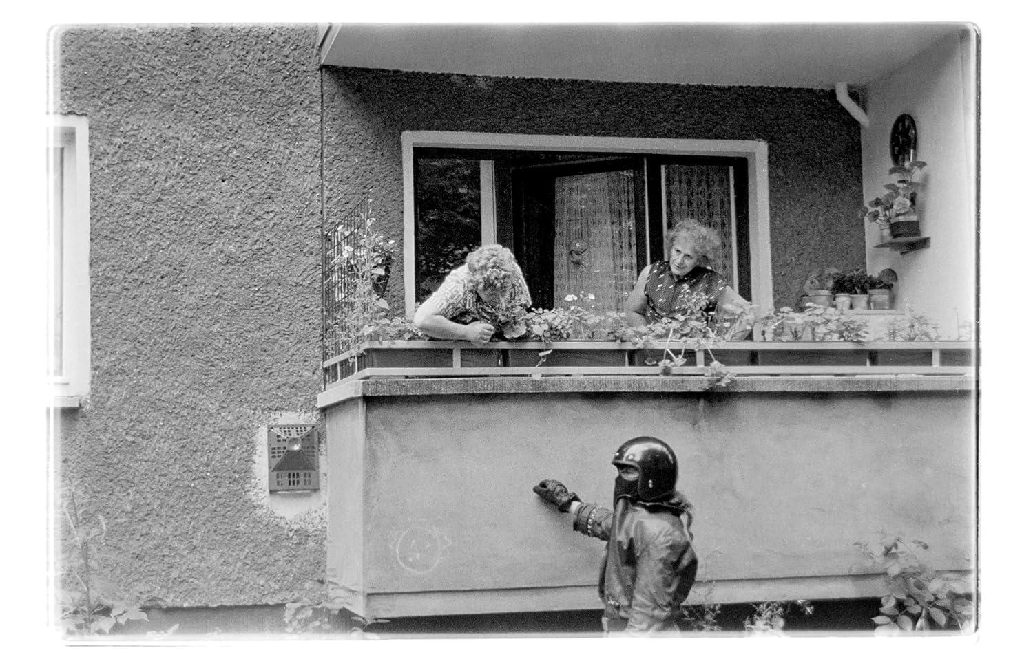 Zwei Frauen auf einem Balkon mit Blumen, eine Person mit Helm zeigt auf die Wand.