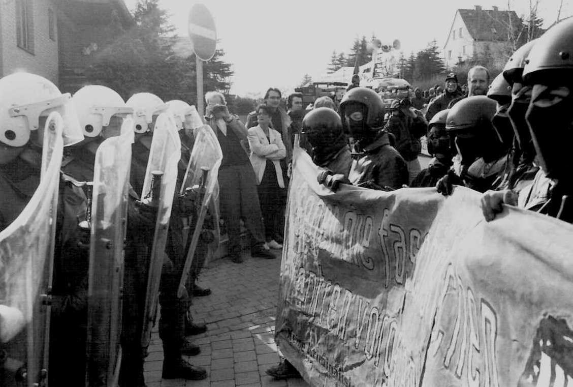 Riot-Polizei mit Schilden steht Protestierenden mit einem Banner gegenüber. Im Hintergrund ein Straßenschild und Gebäude. Schwarzweiß-Foto.