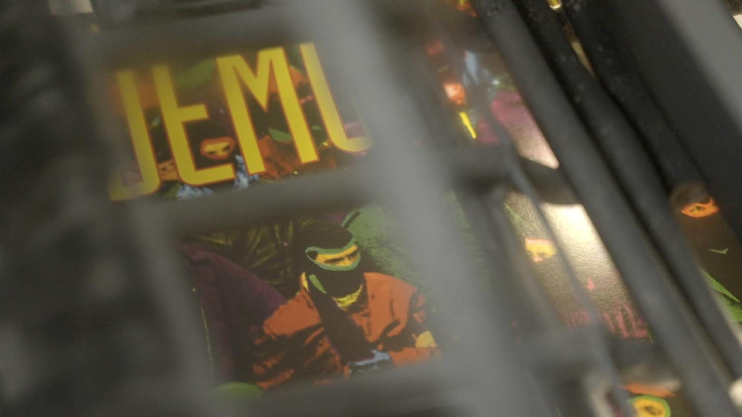 Detail der Flipper-Maschine. Gelber 'JEMU'-Text sichtbar. Eine Person in orangefarbener Jacke und schwarzer Maske ist hinter den Stäben zu sehen.