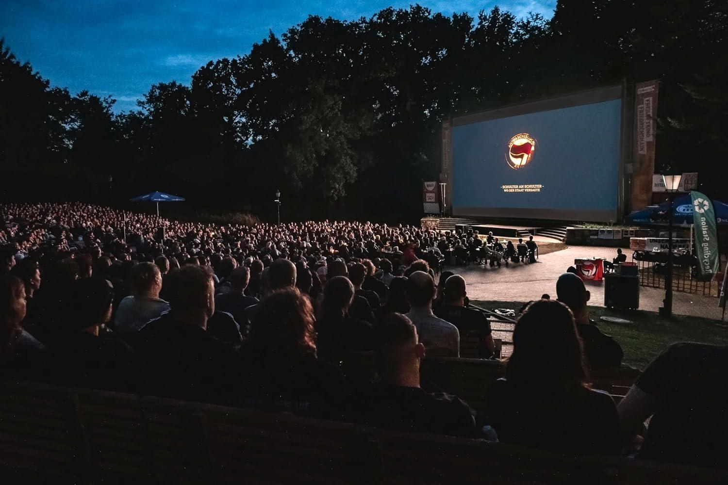 Freiluftkino mit großem Bildschirm, viele Menschen schauen zu. Dunkelblauer Himmel, Bäume im Hintergrund.
