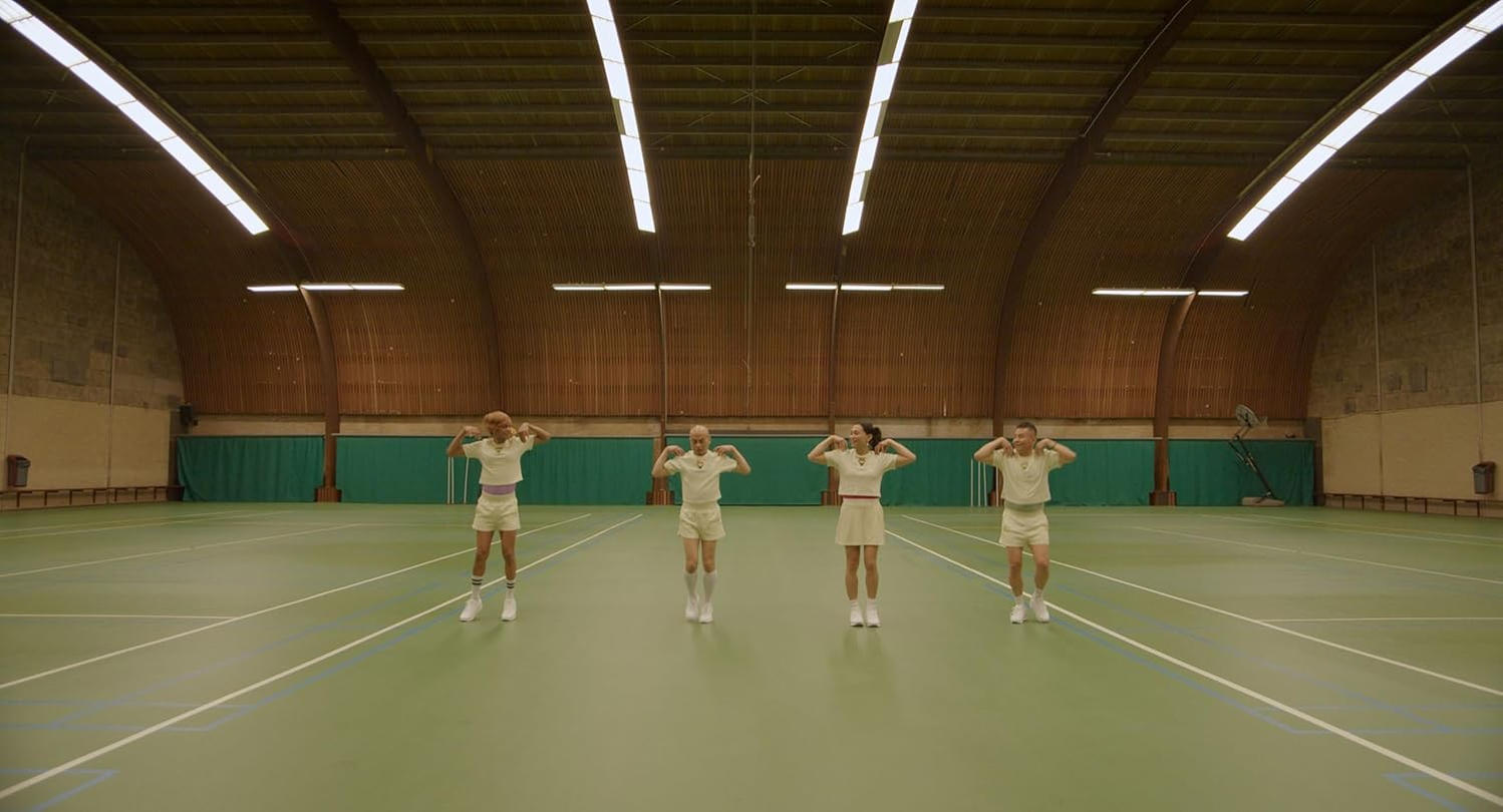 Vier Personen in passenden Outfits stehen auf einem Tennisplatz mit erhobenen Armen. Sie befinden sich in einem großen Innenraum mit Holzverkleidung.