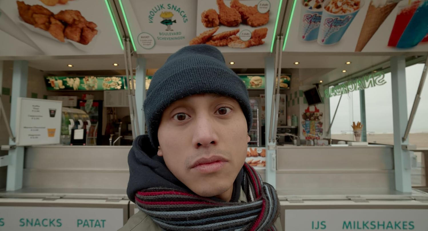 Ein Mann mit Mütze und Schal steht vor einem Imbissstand. Der Stand verkauft Snacks, Pommes, Eis und Milchshakes.