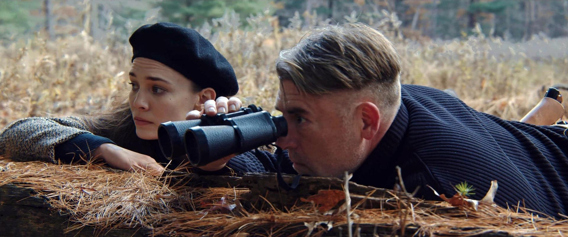 Zwei Personen im Wald, eine blickt durch ein Fernglas. Frau mit Baskenmütze. Laub und Kiefernnadeln im Vordergrund.