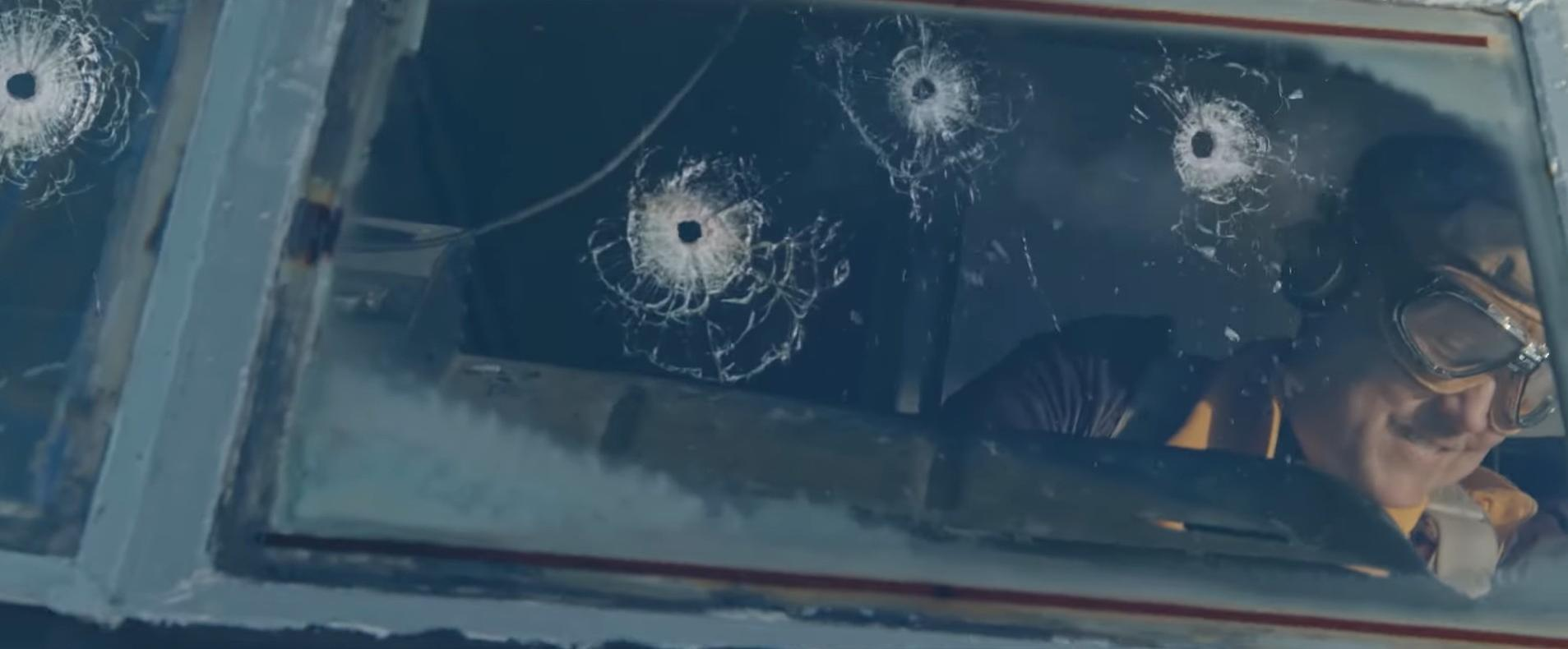 Ein Pilot blickt durch ein zerbrochenes Flugzeugfenster. Einschusslöcher sind im Glas zu sehen. Der Pilot trägt eine Brille und einen gelben Anzug.