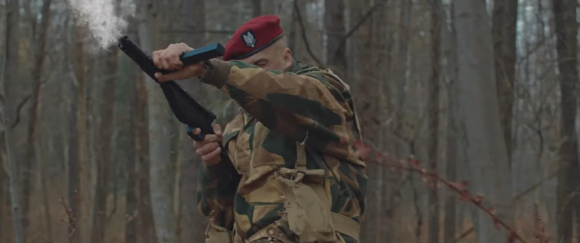 Ein Soldat in Tarnkleidung und rotem Barett schießt in einem Wald mit einer Schrotflinte. Rauch quillt aus dem Lauf.